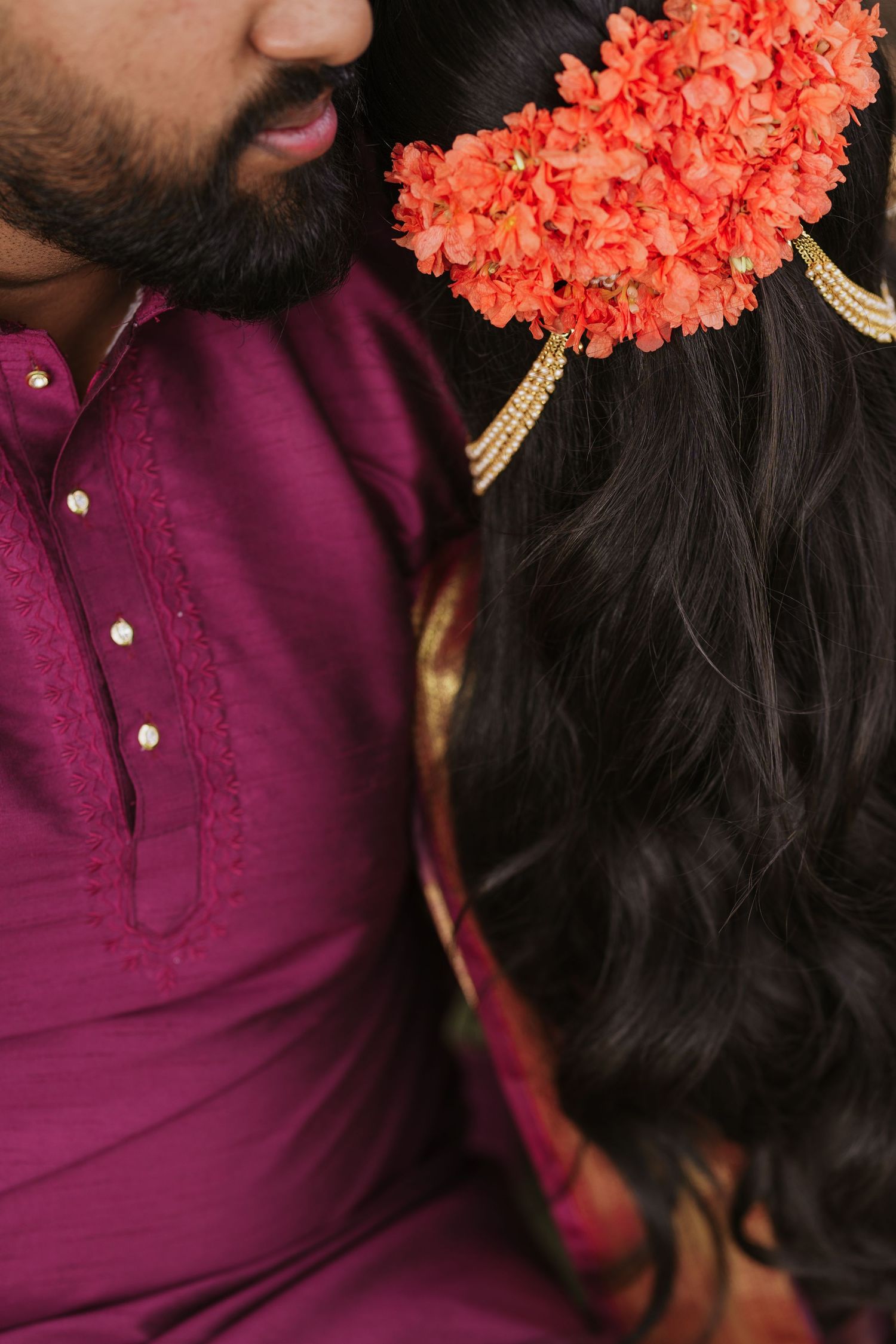 Indian couple in traditional attire, man in purple kurta beside woman with red floral hair accessory and gold jewelry.