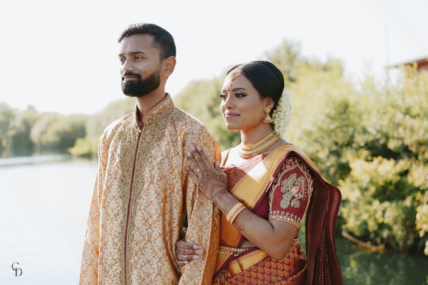 A couple in traditional Indian wedding attire poses outdoors near a waterfront.