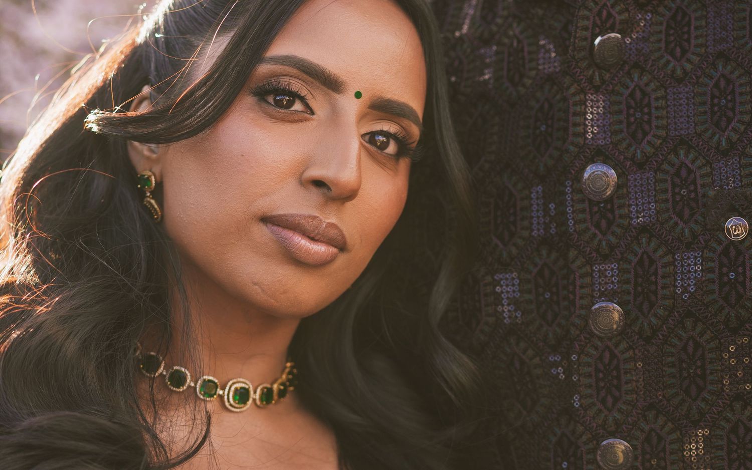 A young South Asian woman wearing a gold necklace and traditional attire poses elegantly against a dark backdrop.
