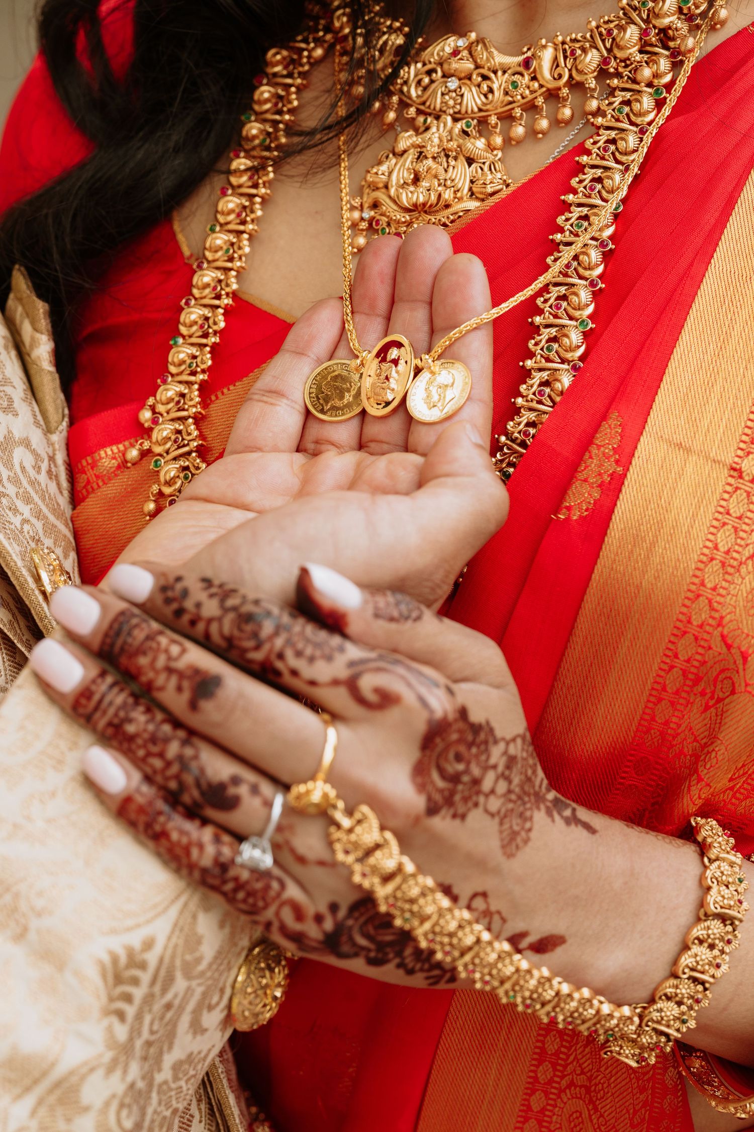 Indian bride in red saree displaying gold coin pendants on henna-adorned hands with traditional gold jewelry.