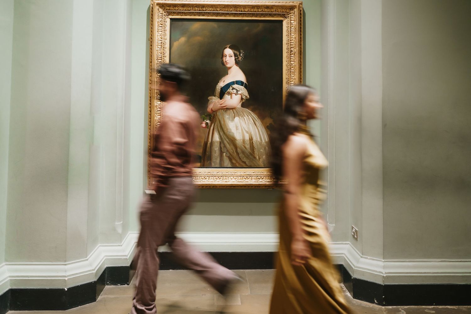 Museum visitors move quickly past artwork in a blurred motion through an elegant gallery space with framed paintings.