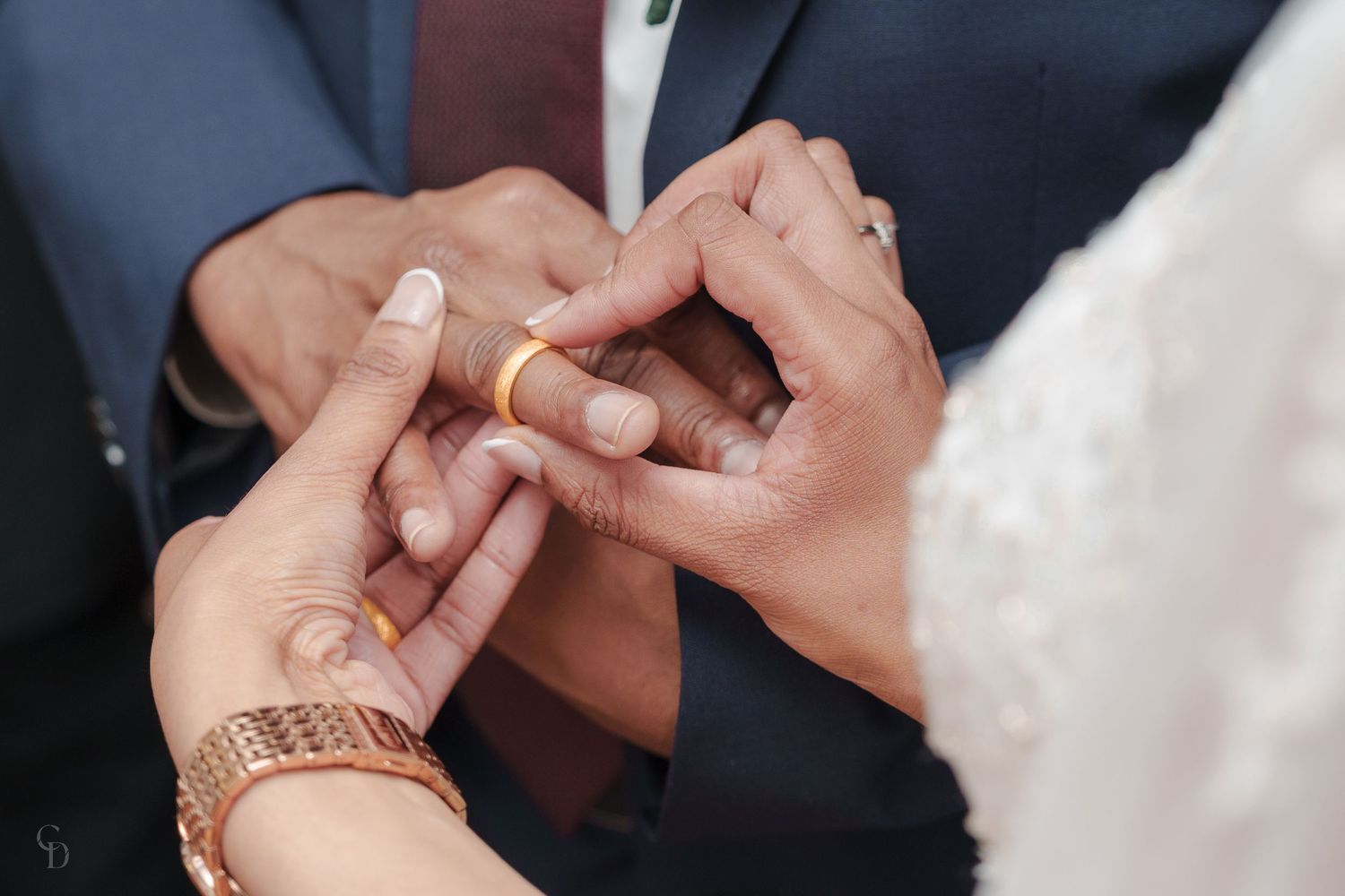 Detailed shot of hands exchanging wedding rings with gold jewelry and white lace details.