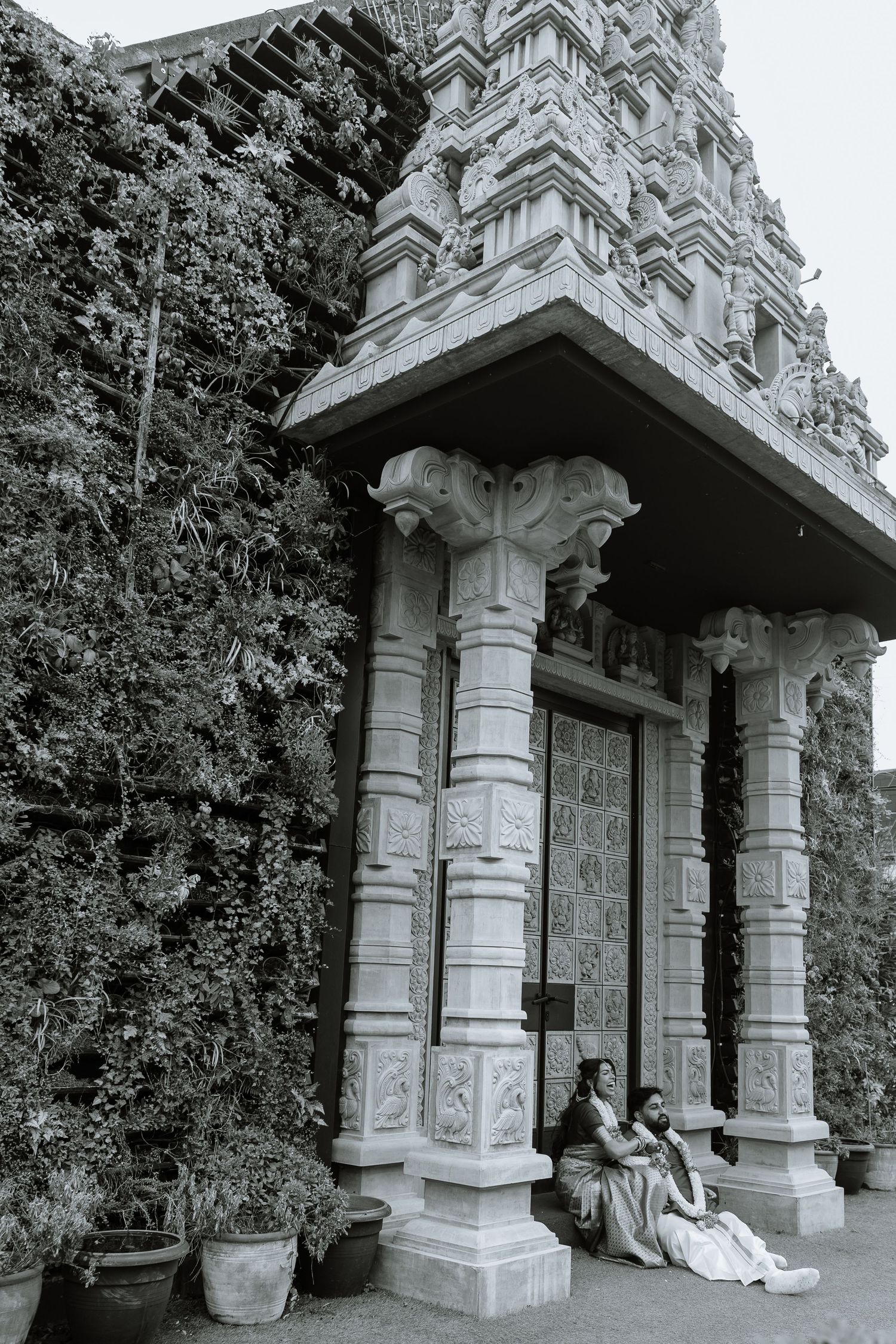 Black and white photo of two people sitting beside ornate carved columns at a Hindu temple entrance with lush greenery.