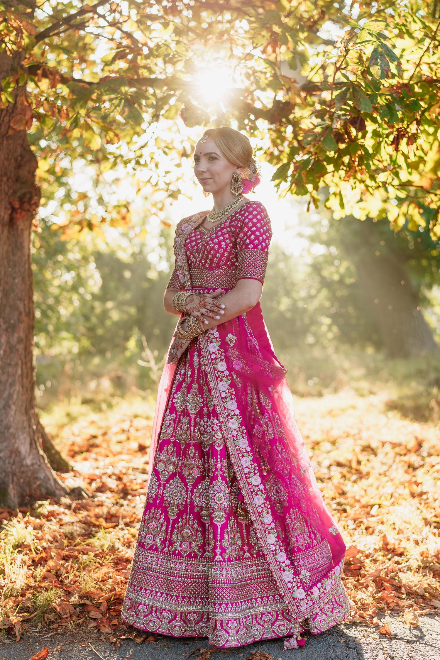 A traditional pink and gold lehenga sparkles in autumn sunlight under a tree with falling leaves creating a magical atmosphere.