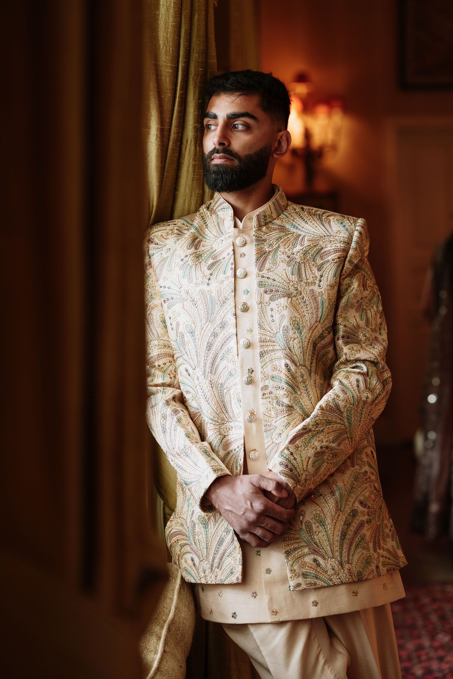 A man wearing an ornate cream and gold embroidered sherwani suit stands in front of a warm-lit curtained backdrop.