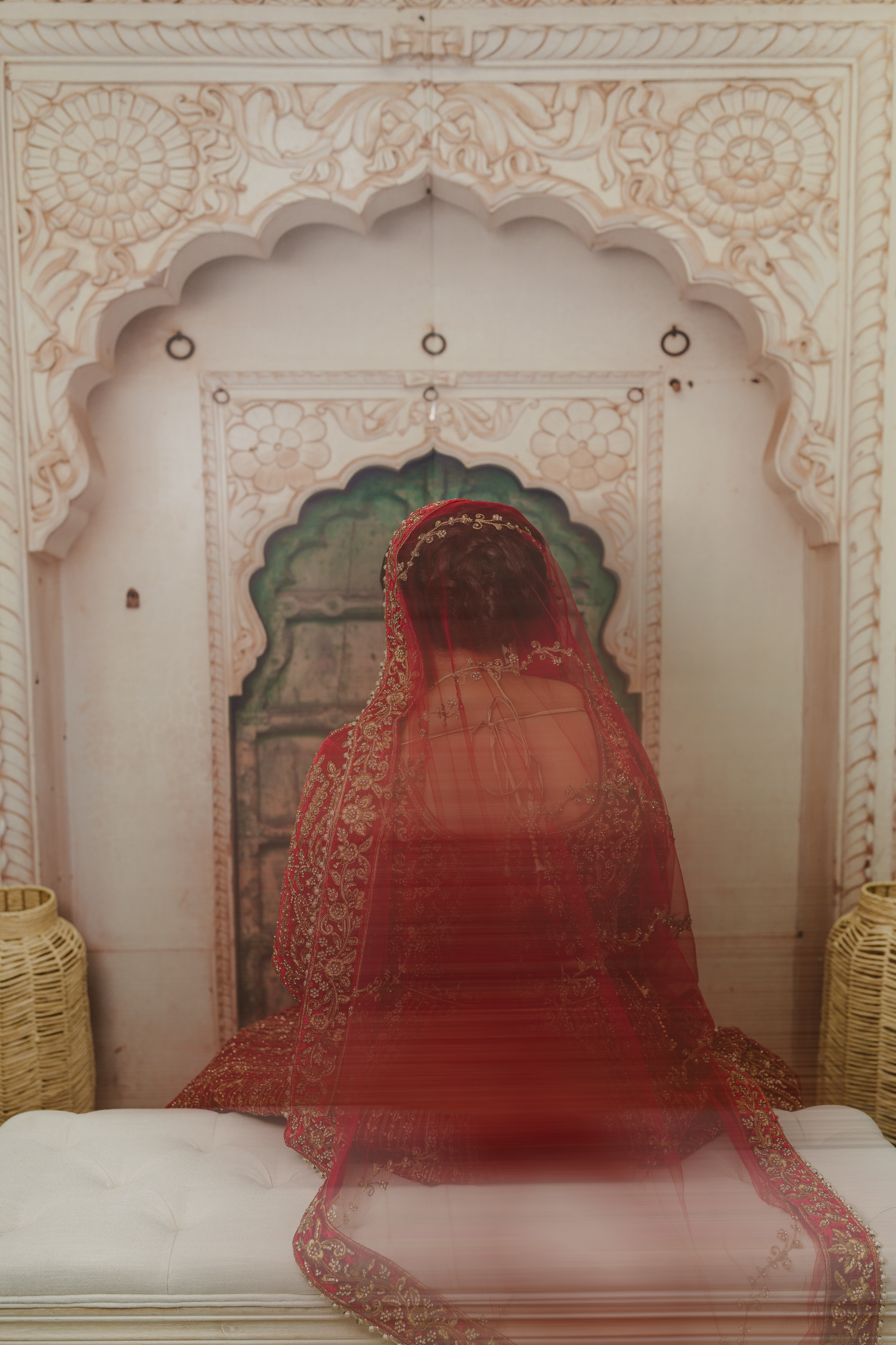Indian bride in red bridal lehenga and dupatta veil sitting in ornate white Rajasthani arch alcove.