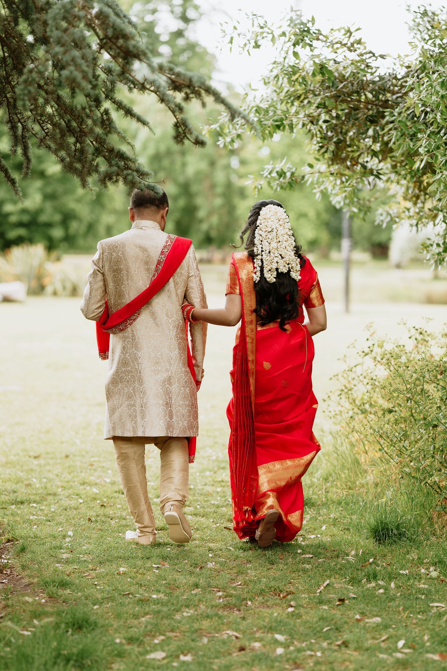Couple walks hand in hand through a sun-dappled garden path wearing traditional South Asian wedding attire in beige and red.
