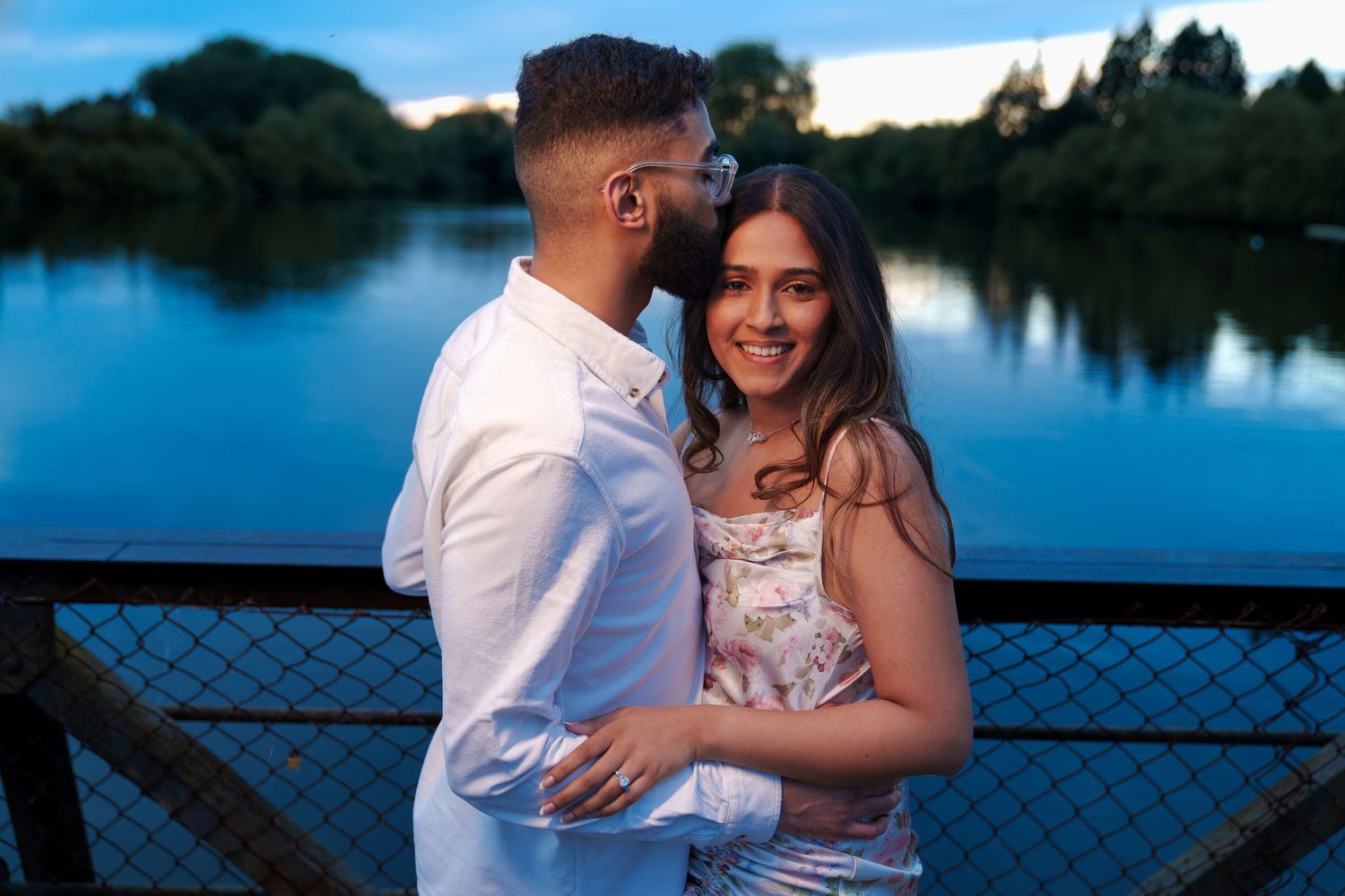 Joyful couple laughing while embracing on bridge with reflective lake behind them.