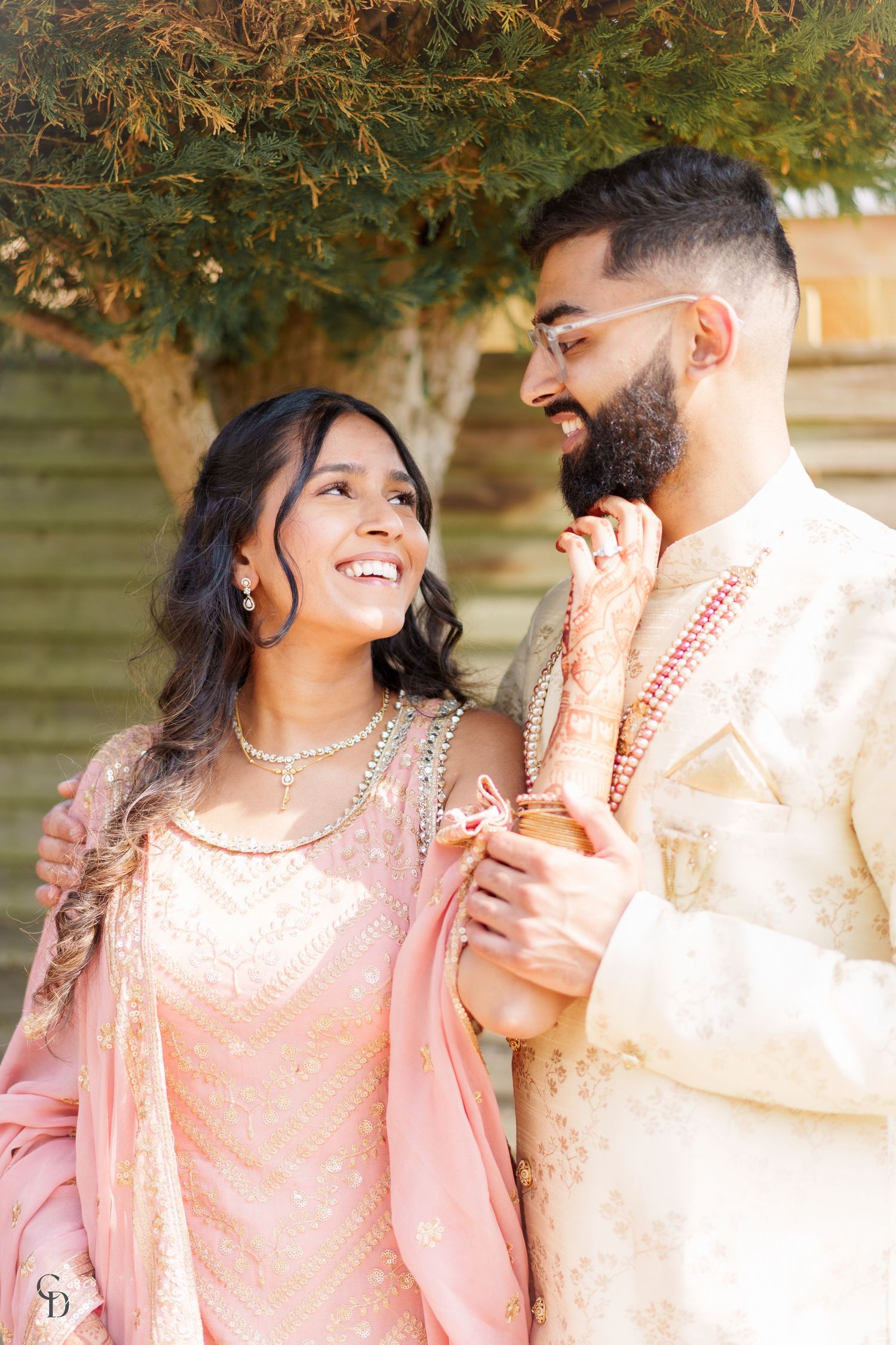 Couple in traditional South Asian attire sharing joyful moment under tree.
