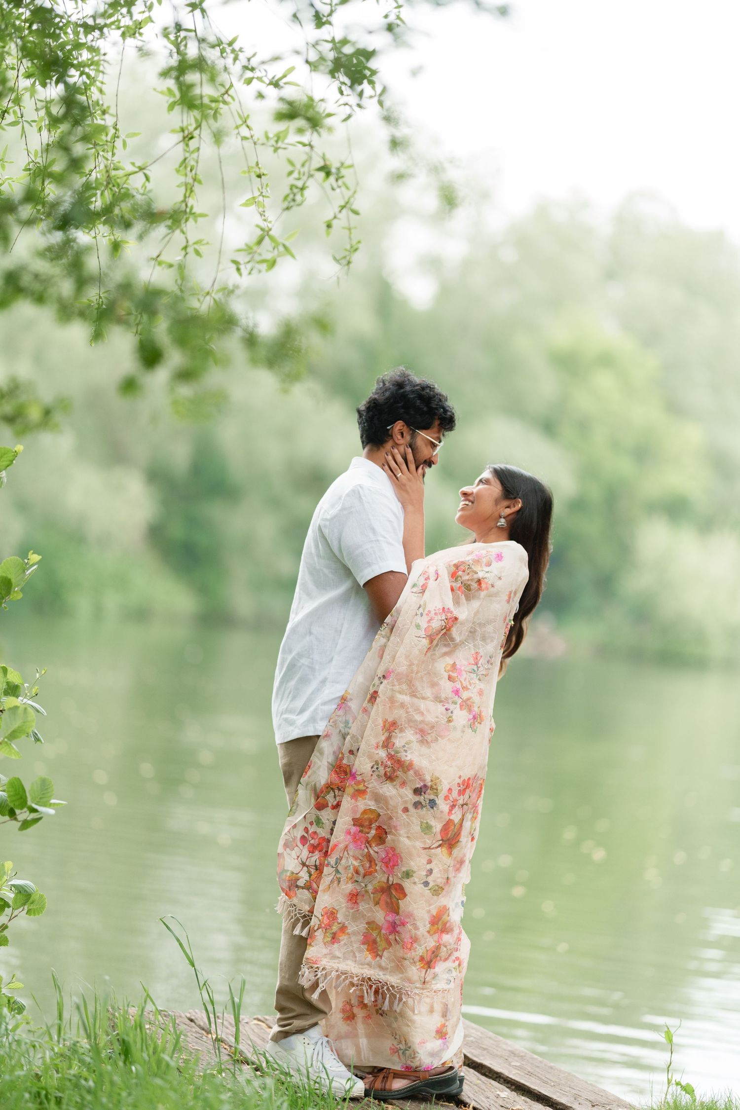 A romantic couple embraces by a serene lake, with the woman wearing a floral peach sari against a dreamy natural backdrop.