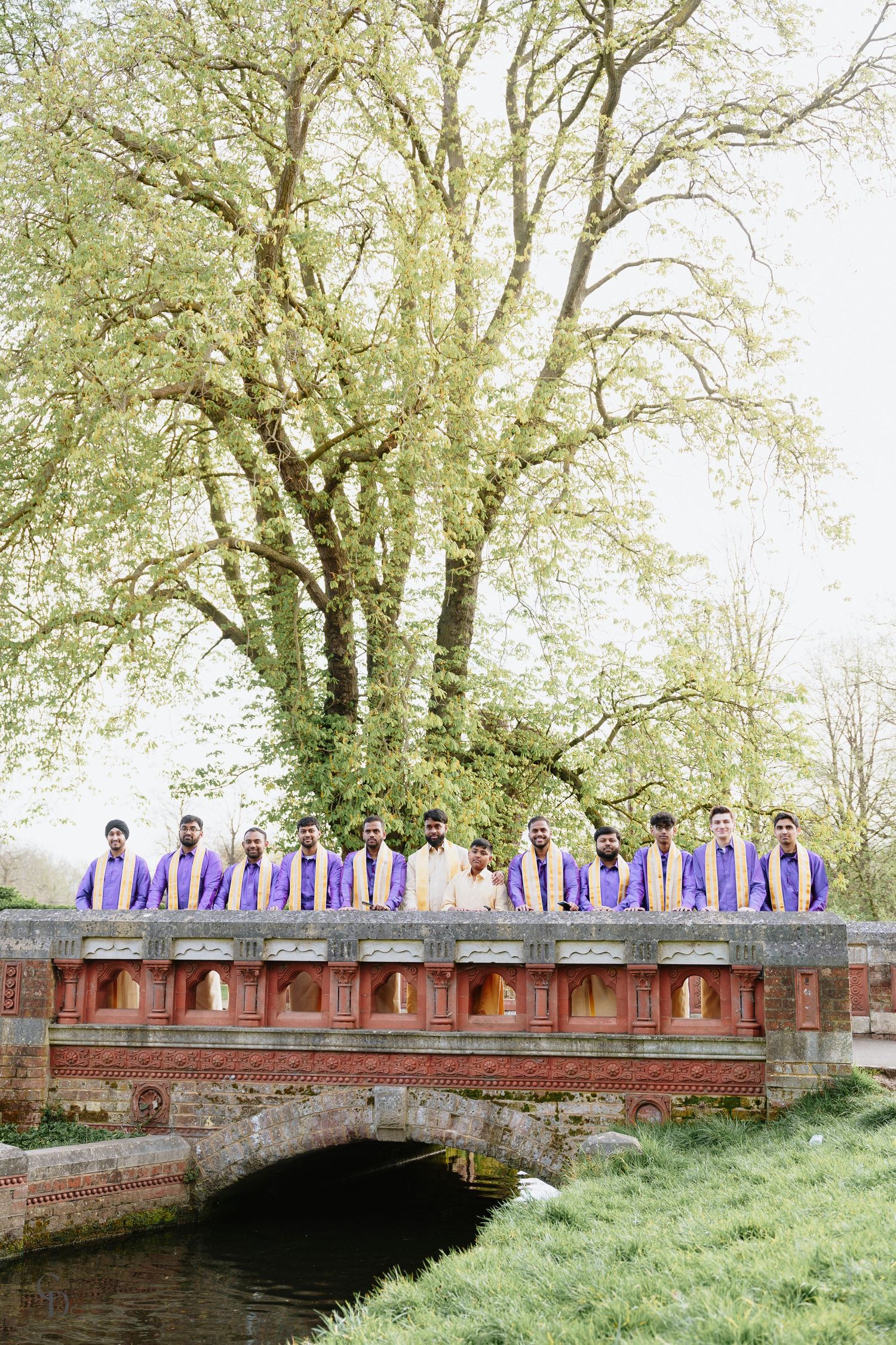 Wedding party in purple attire standing on stone bridge against backdrop of spring trees.
