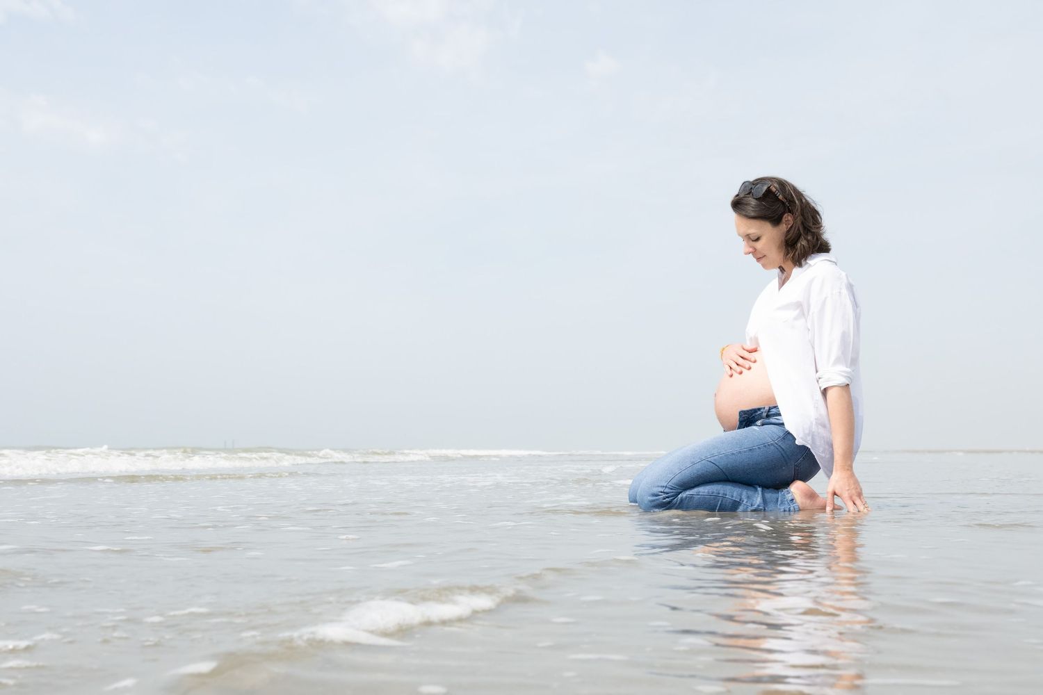 Pregnant woman kneeling in shallow ocean waves on a beach, cradling her baby bump while wearing jeans and a white shirt.
