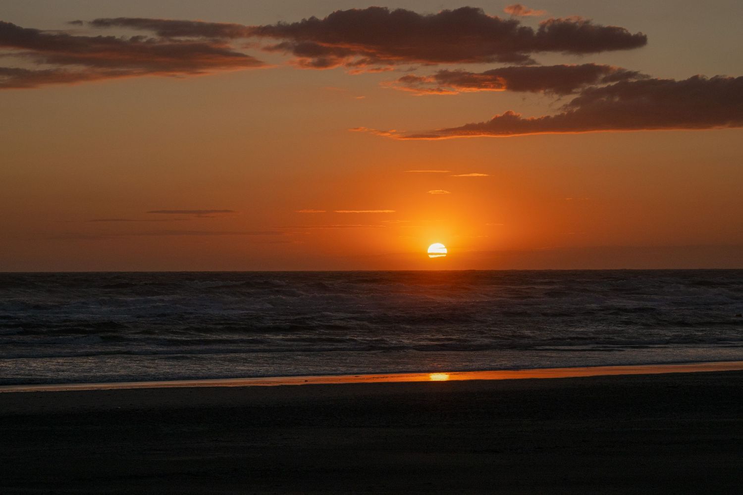 Golden sunset over ocean horizon with vibrant orange sky, scattered clouds, and calm waves on a serene beach.