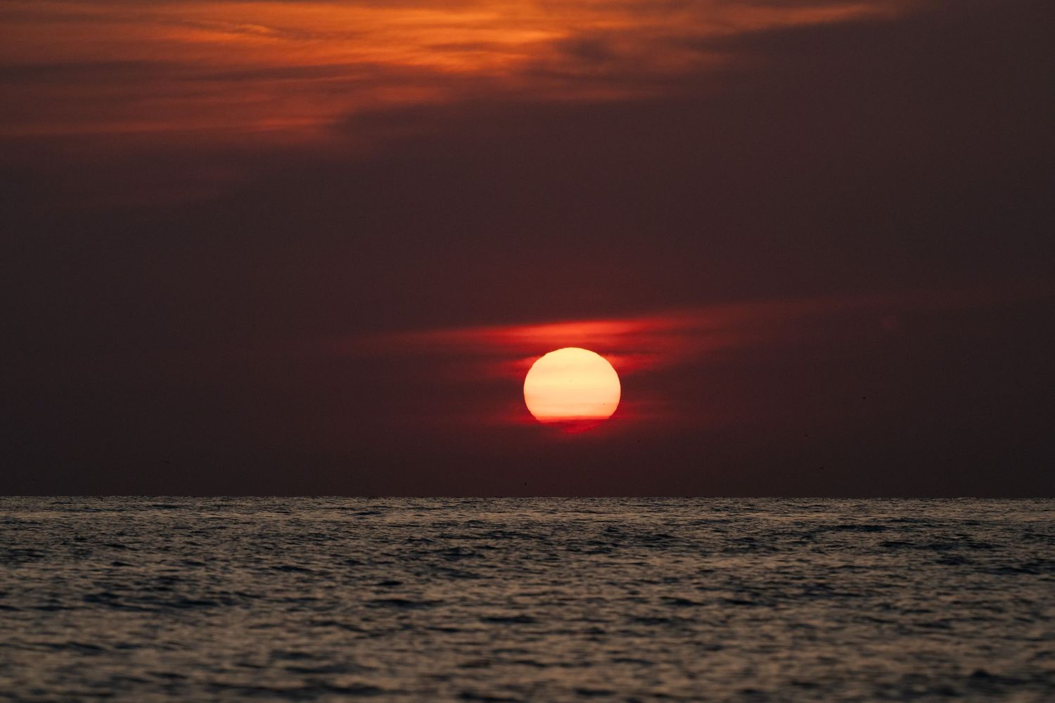 Dramatic red sunset over calm ocean waters with large sun setting on the horizon under dark cloudy skies.