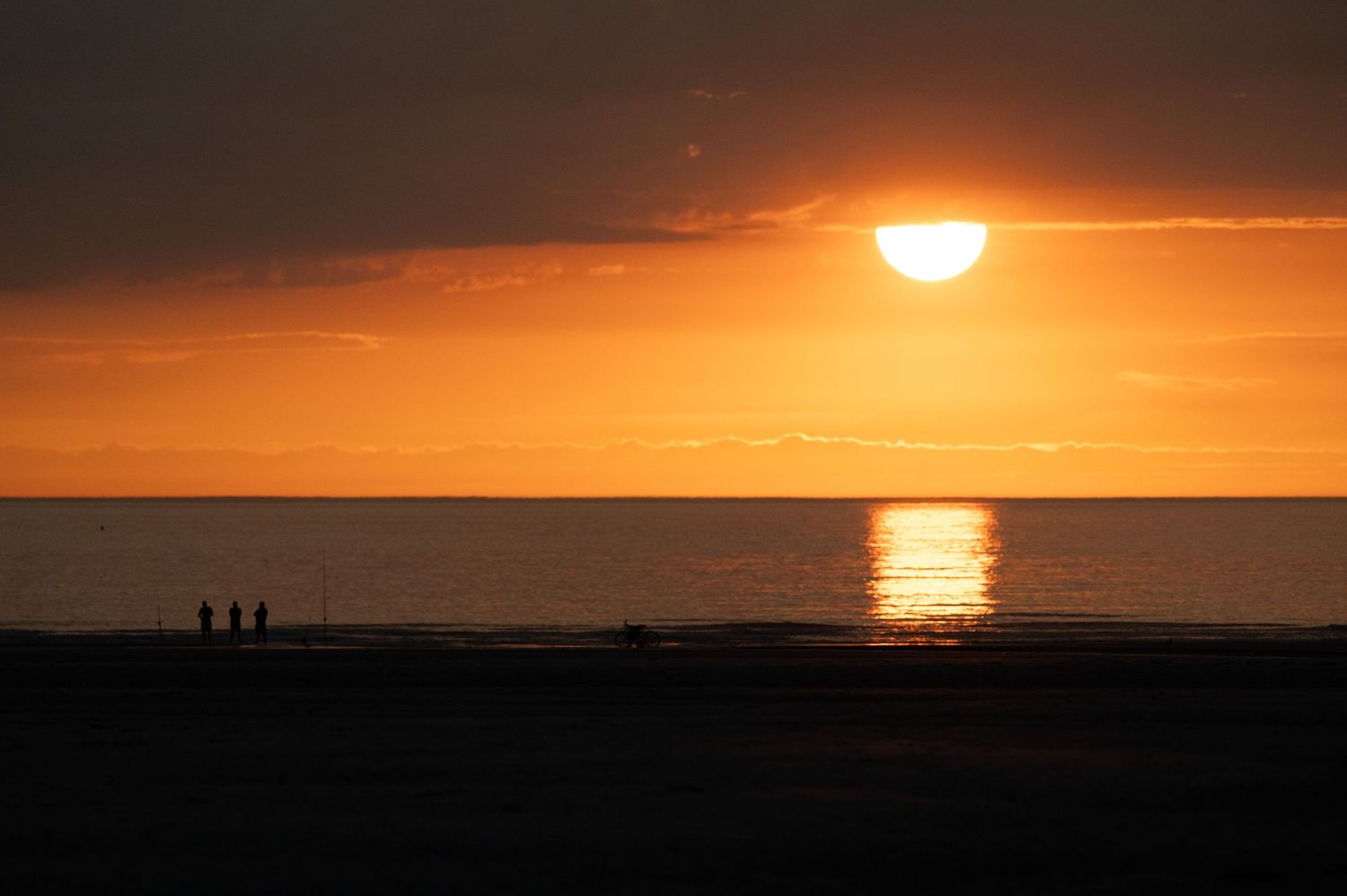 Silhouettes of people on a beach at golden sunset with sun reflecting on calm ocean waters.