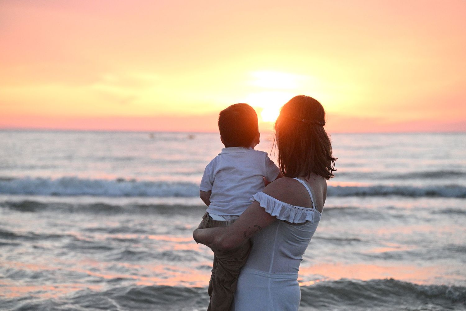 A mother holds her young child while watching a beautiful golden sunset over the ocean waves at the beach.