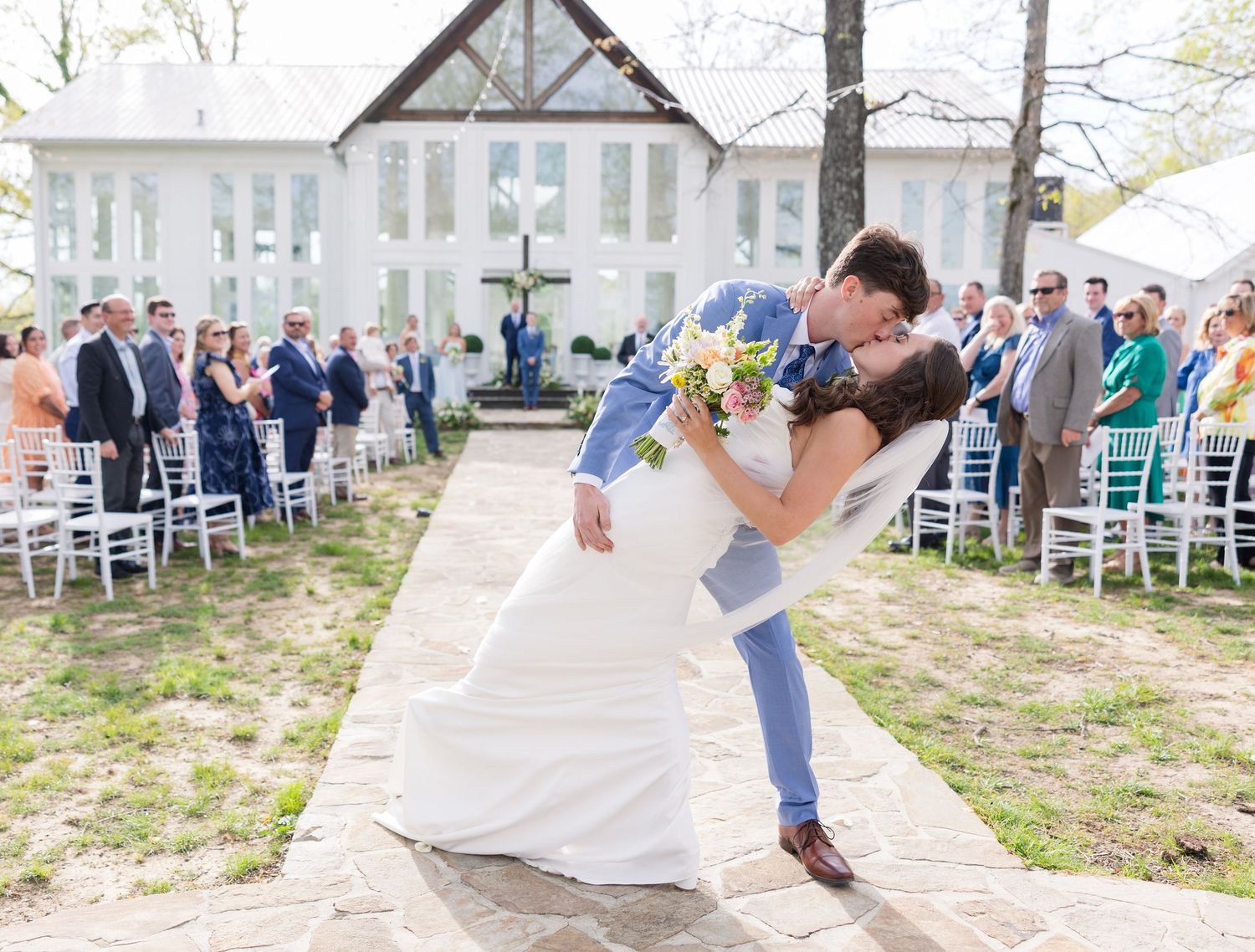 Wedding guests gather outdoors near a gazebo in spring with rows of white chairs set up for a ceremony.