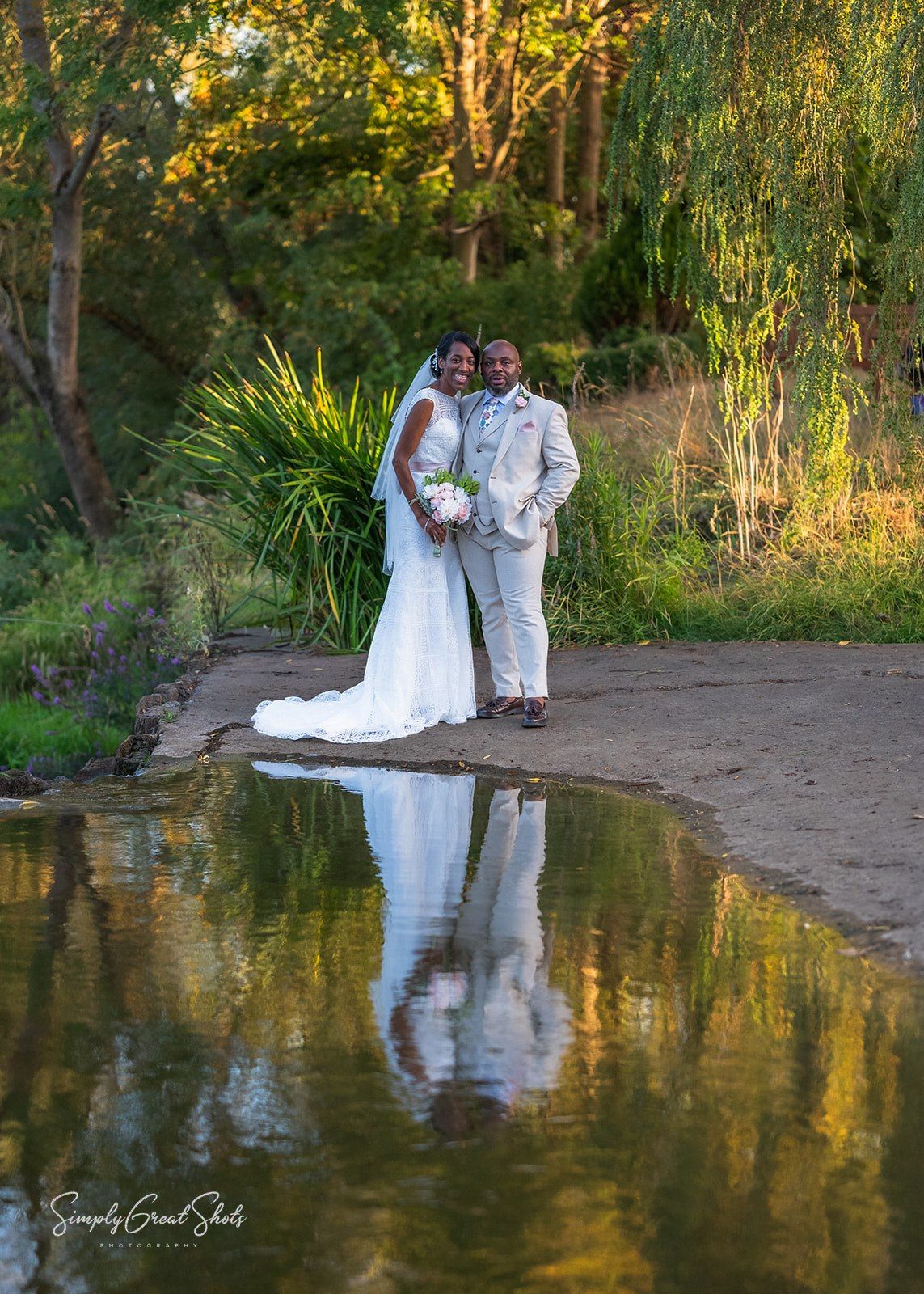 Wedding couple poses together near a tranquil pond at sunset with golden light reflecting on the water's surface at Stratford Upon Avon.