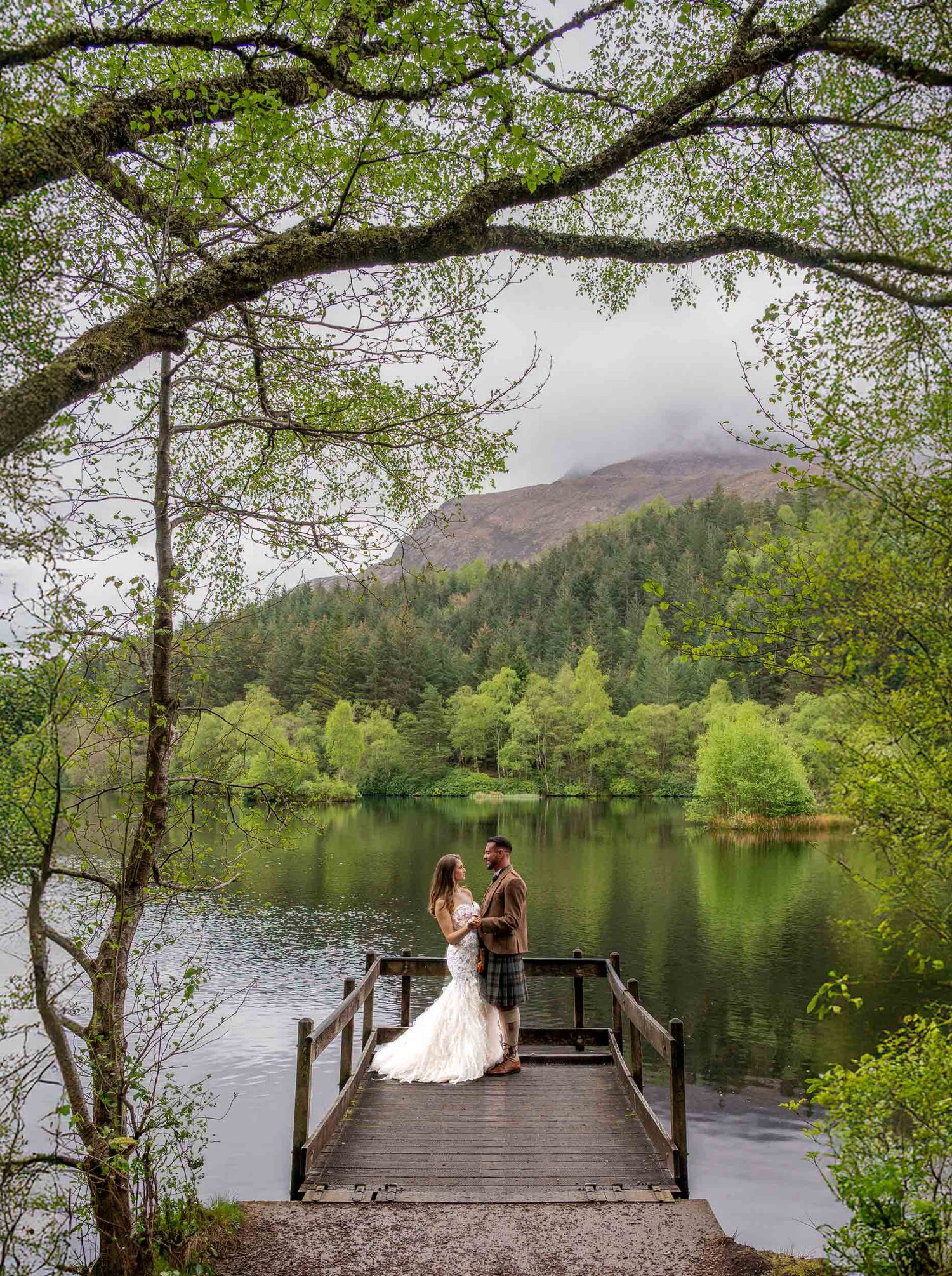 A couple stands on a wooden jetty overlooking a tranquil lake surrounded by lush green forest and mountains in Scotland.
