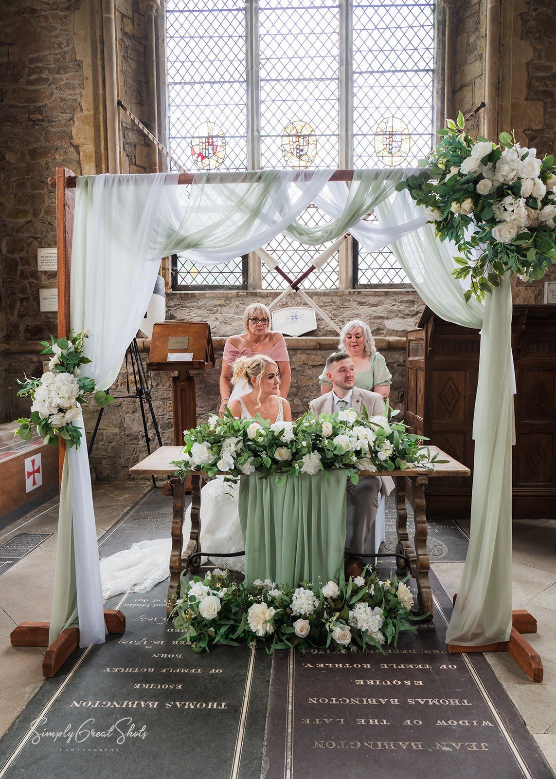 Wedding ceremony in a historic stone chapel with elegant white floral arrangements and draped fabric decorations.