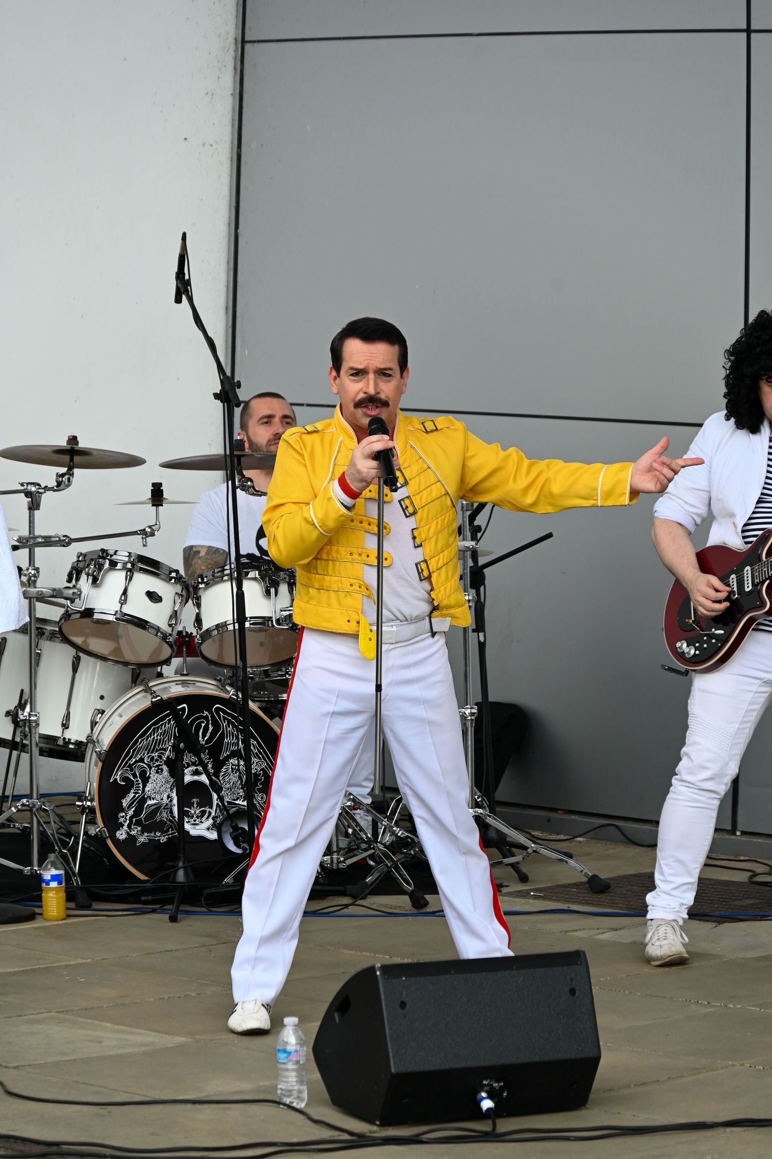 A Freddie Mercury tribute singer in a yellow jacket and white pants sings on stage with a drum kit and guitarist visible in the background.