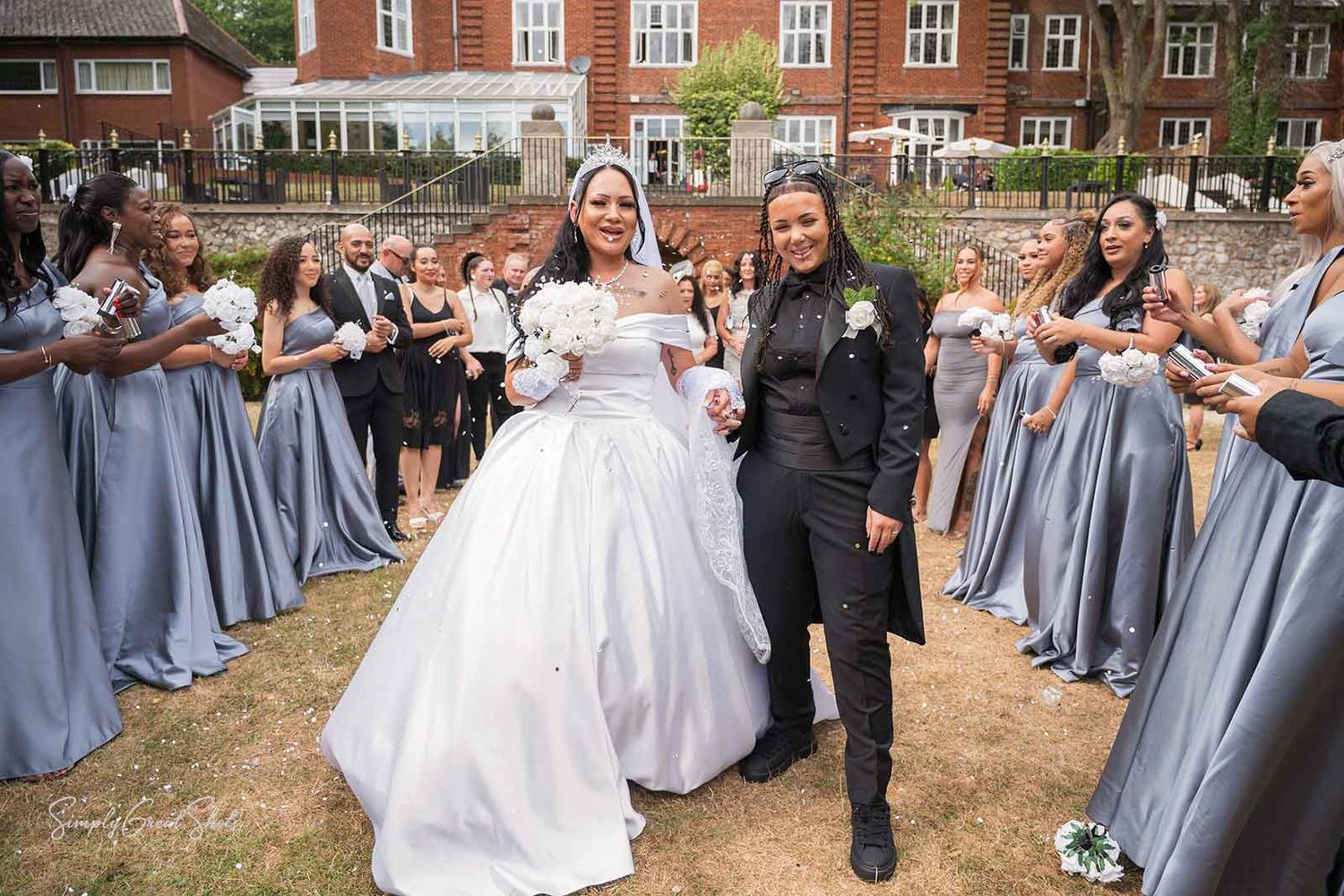 Wedding party wearing pale gray bridesmaid dresses forms a line outdoors with a white-gowned figure in focus.