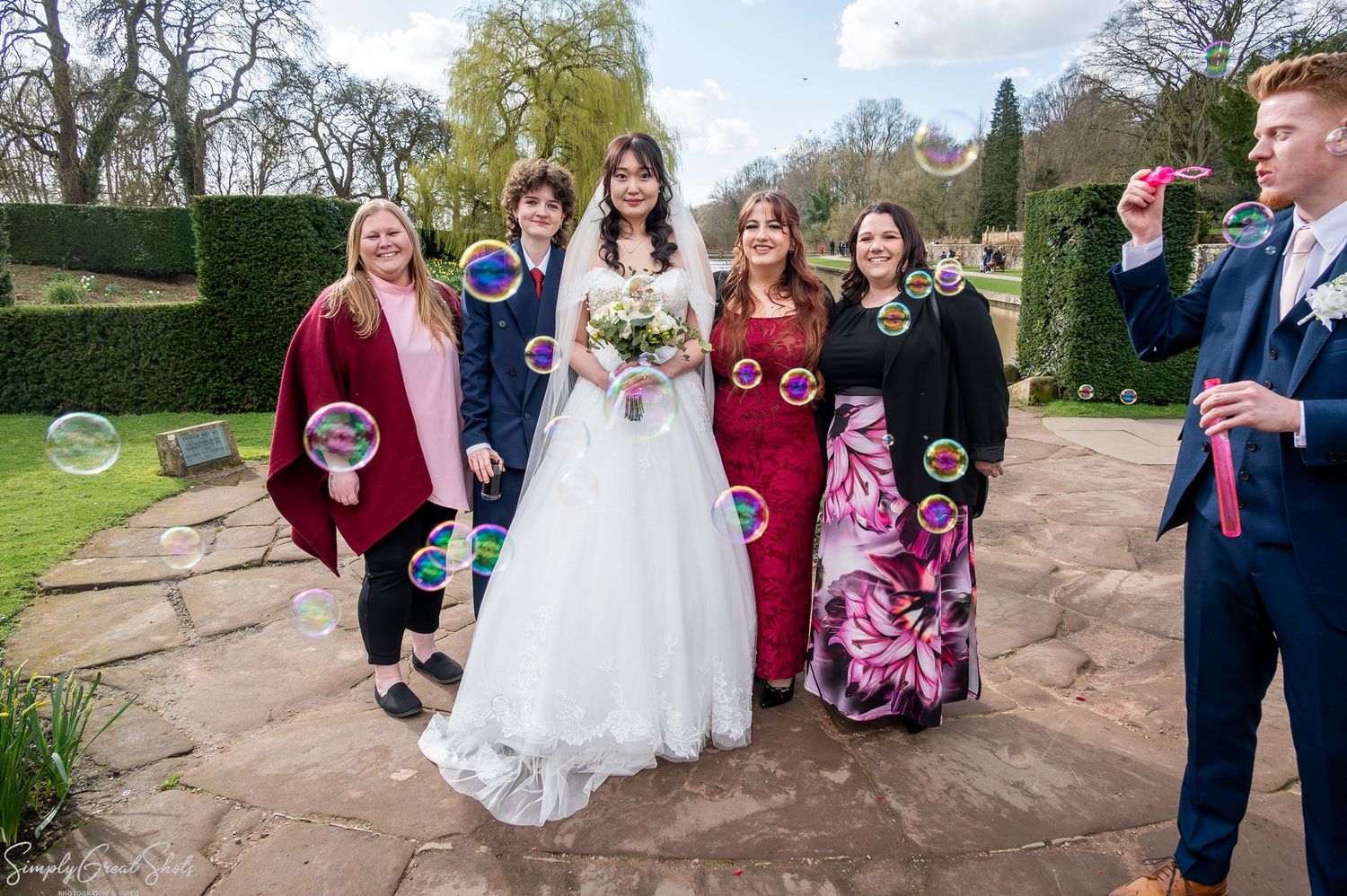 Bride Minky and guests laughing in Coombe Abbey gardens as a groomsman blows bubbles around them.