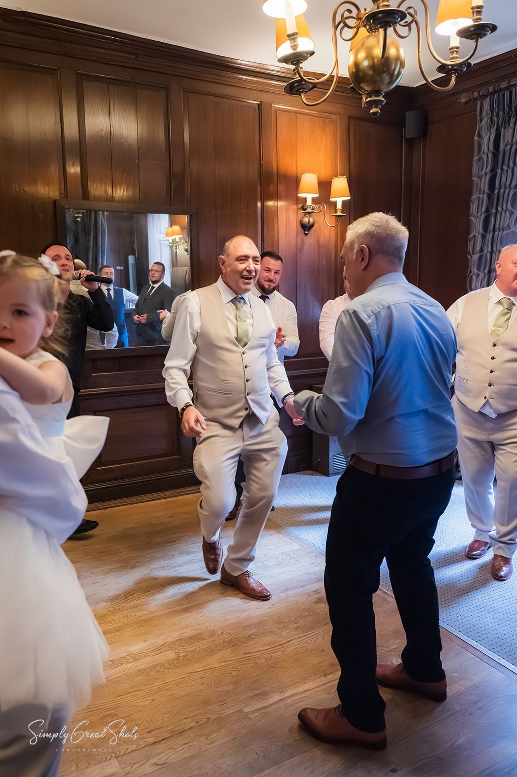 Wedding guests dancing and socialising in an elegant wood-panelled room with warm lighting from ornate chandeliers.