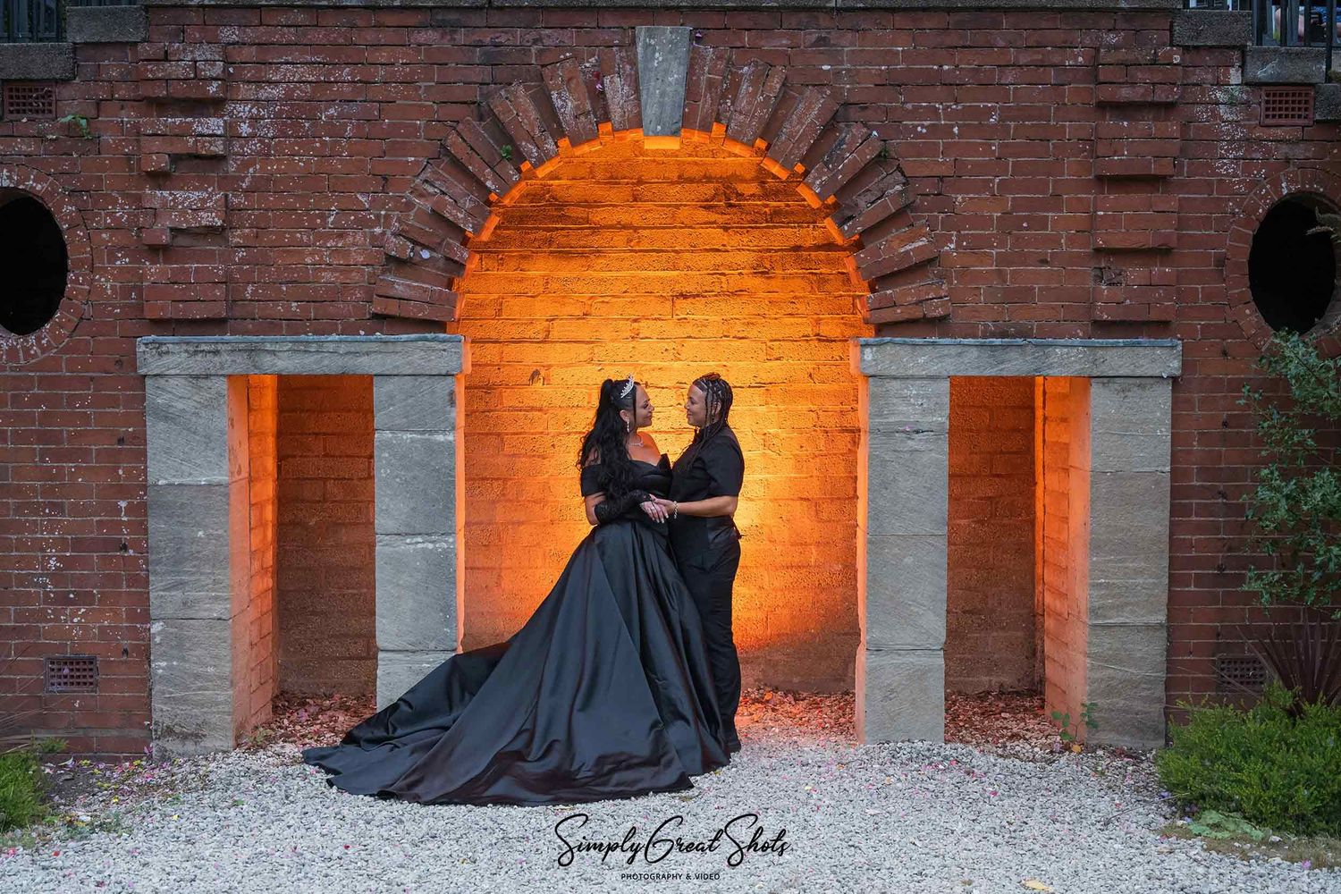Two brides in black clothing pose in front of golden light in an archway at Southcrest Manor Hotel.