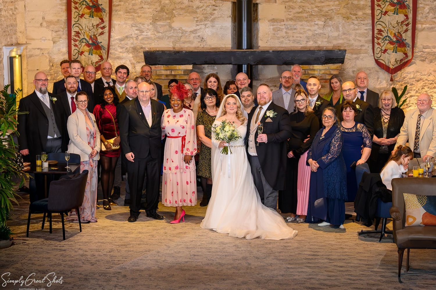 Large wedding party group photograph in historic stone hall with heraldic banners at voco Oxford Thames