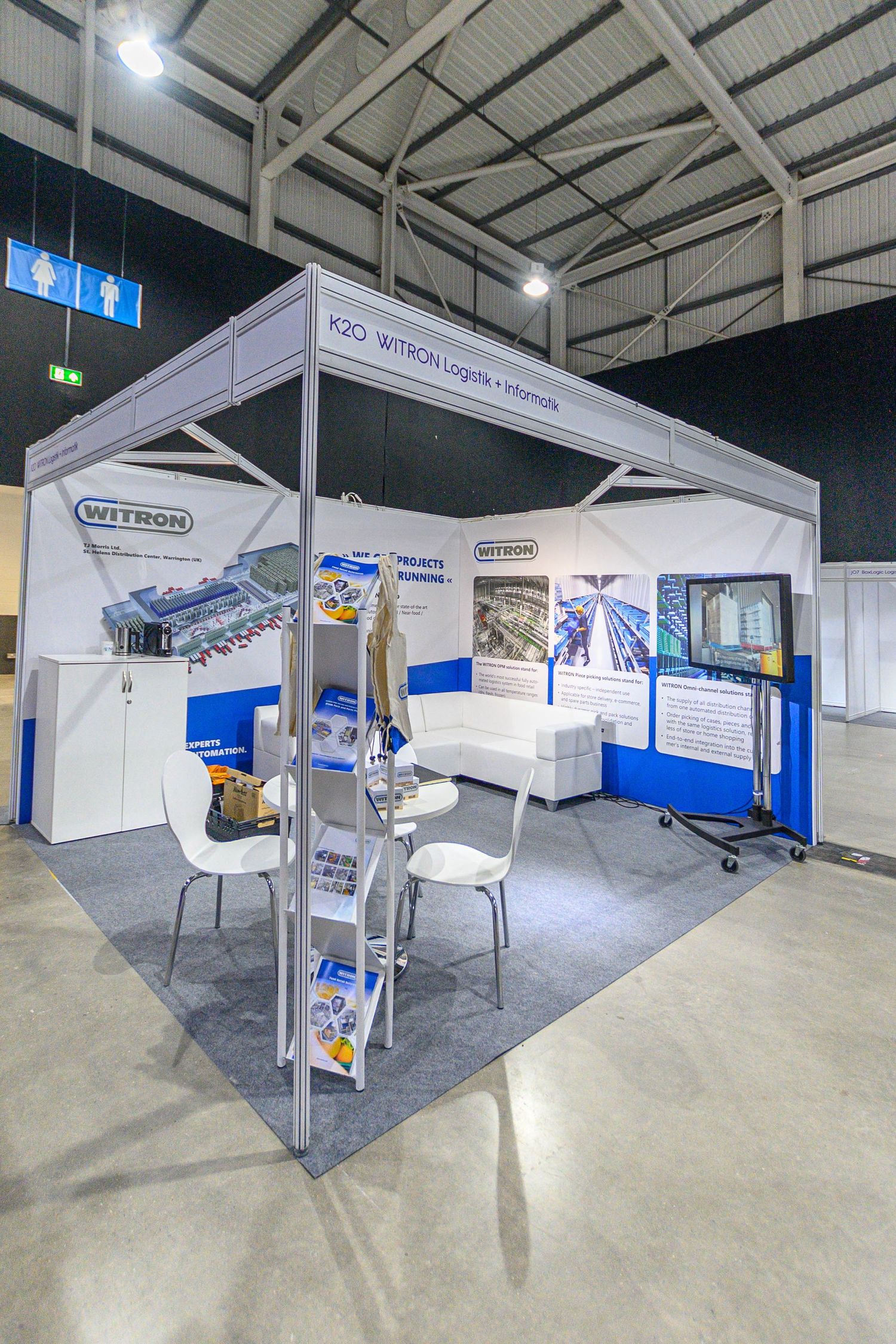Trade show booth displaying industrial equipment with white and blue branding elements and product displays under LED lighting.