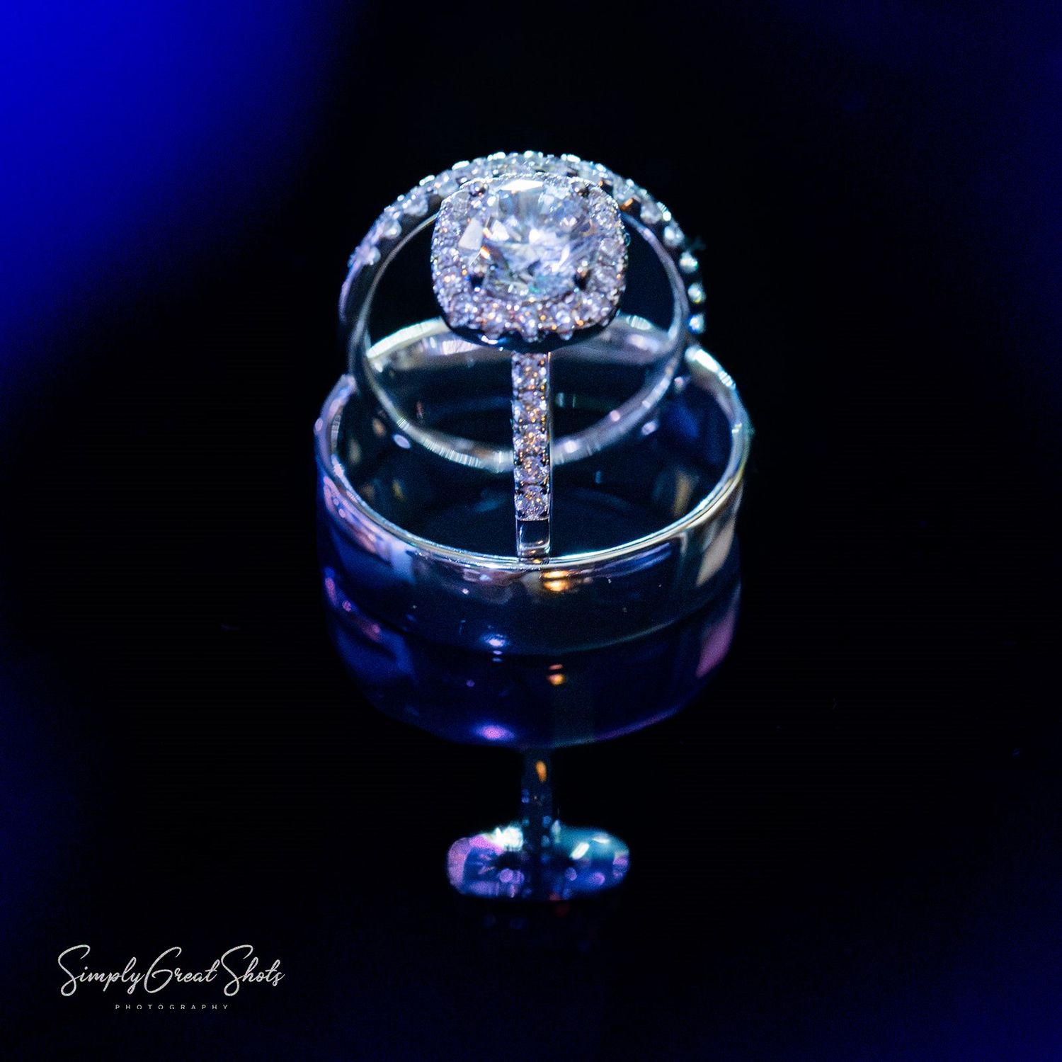 Close-up photo showing a diamond engagement ring and wedding bands on a blue background with dramatic lighting and reflections.