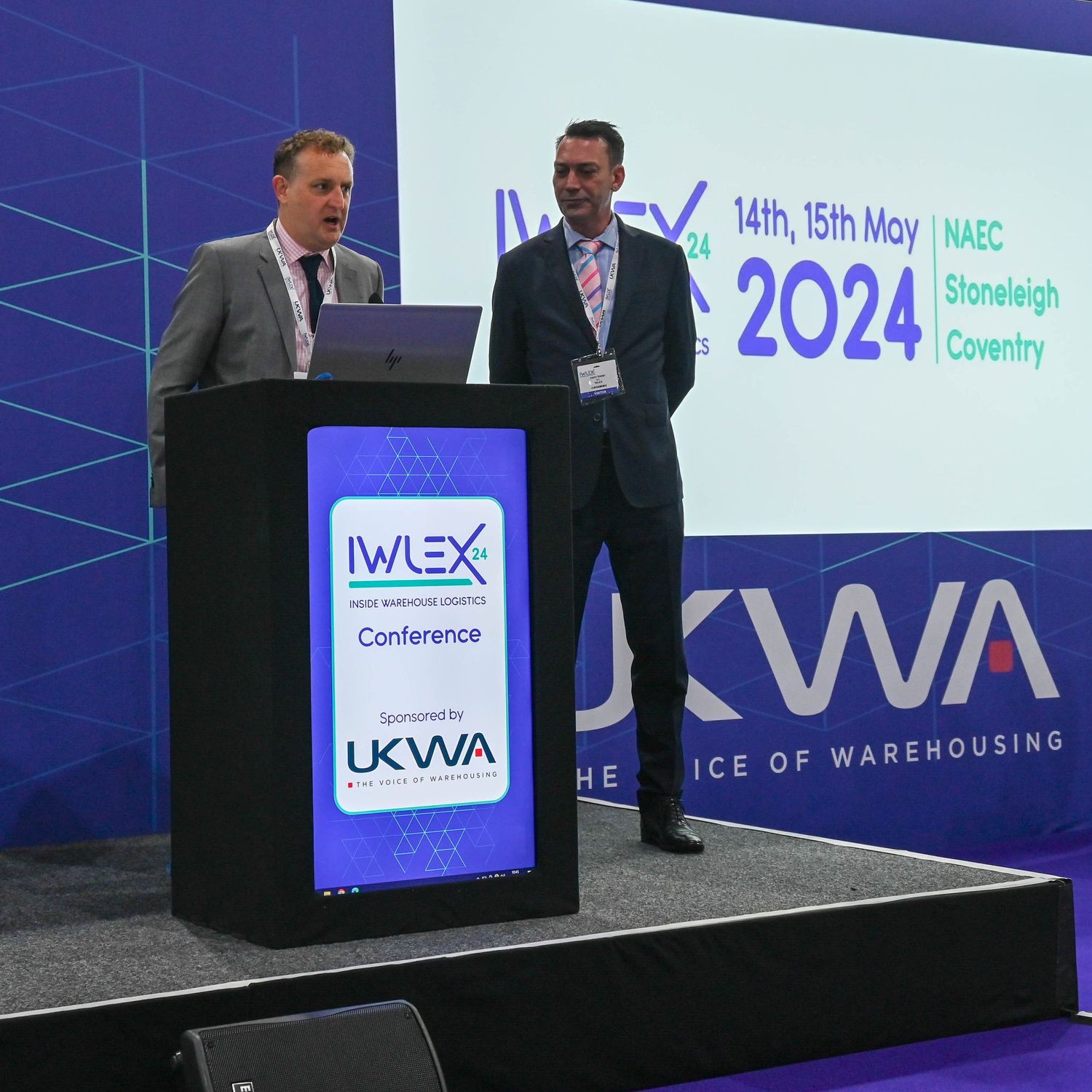 Two presenters stand at a podium during the WLEX Conference with blue branding and signage visible in 2024.