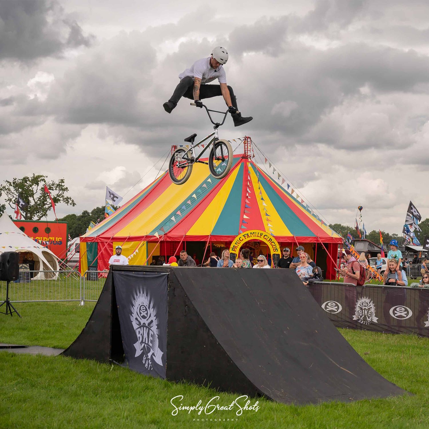 BMX rider performs aerial stunt jump over a black ramp with a colourful circus tent in the background at an outdoor festival.