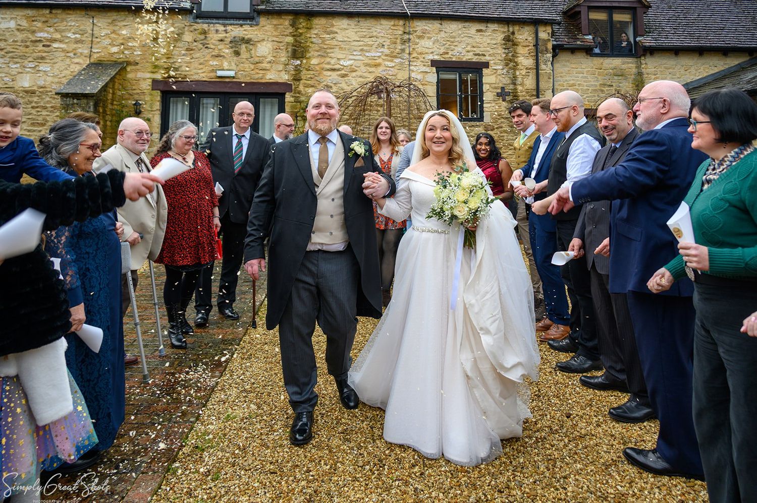 Bride and groom walking through confetti shower with wedding guests lining gravel courtyard at voco Oxford Thames venue