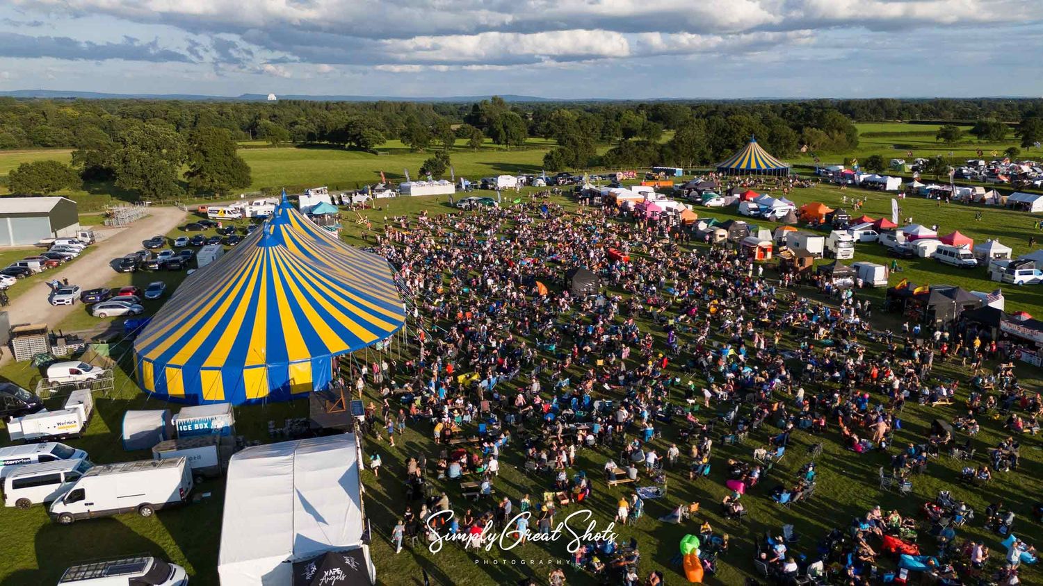 Aerial view of a large outdoor festival with a blue and yellow-striped tent and a massive crowd gathered on a green field.