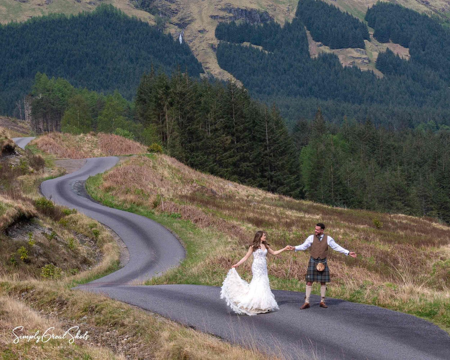 A winding mountain road curves through scenic hills with evergreen trees in the background and golden slopes.