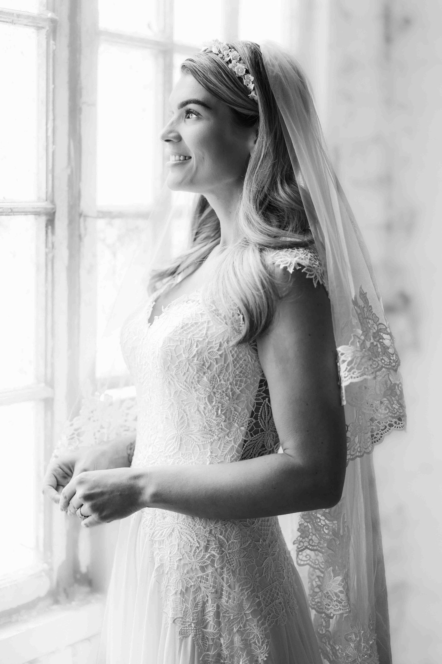 A beautiful bride with a white laced dress and long veil looks out of a window