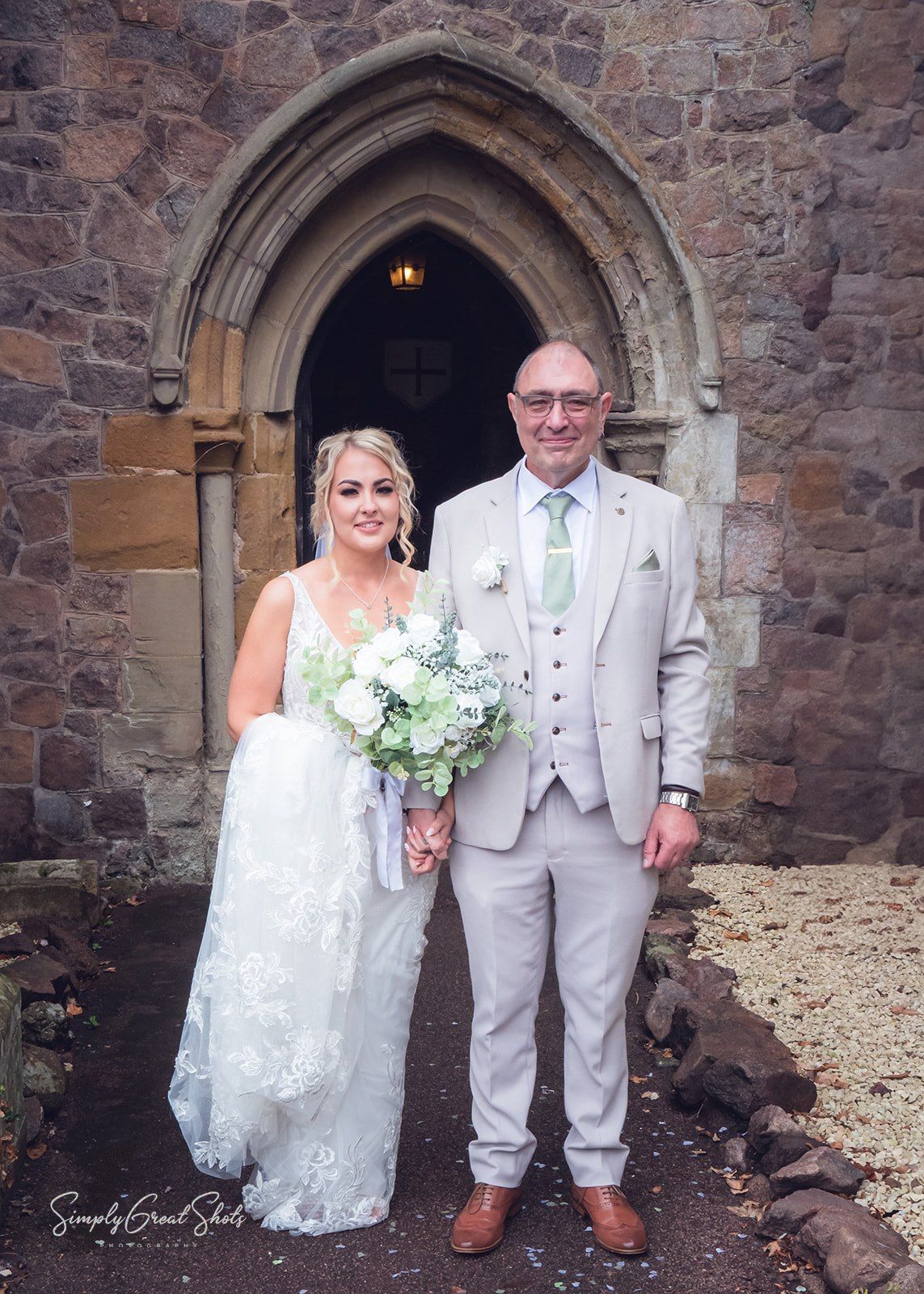 The father of the bride & bride stand in front of a stone church archway at Rothley Temple. The bride is holding a white and green bouquet.