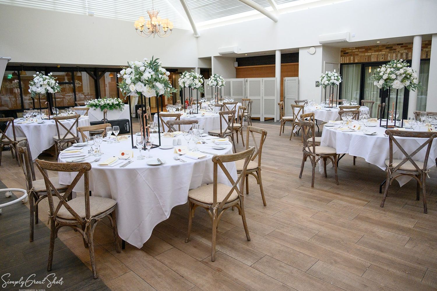 Elegant wedding reception room setup with white linens, tall floral centerpieces, and cross-back chairs at voco Oxford Thames