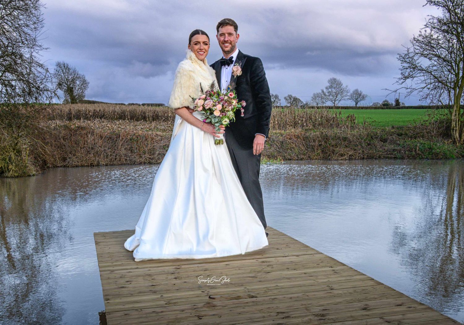 Bride and groom posing on a wooden dock by a lagoon on their wedding day, with cloudy skies and green fields behind them.