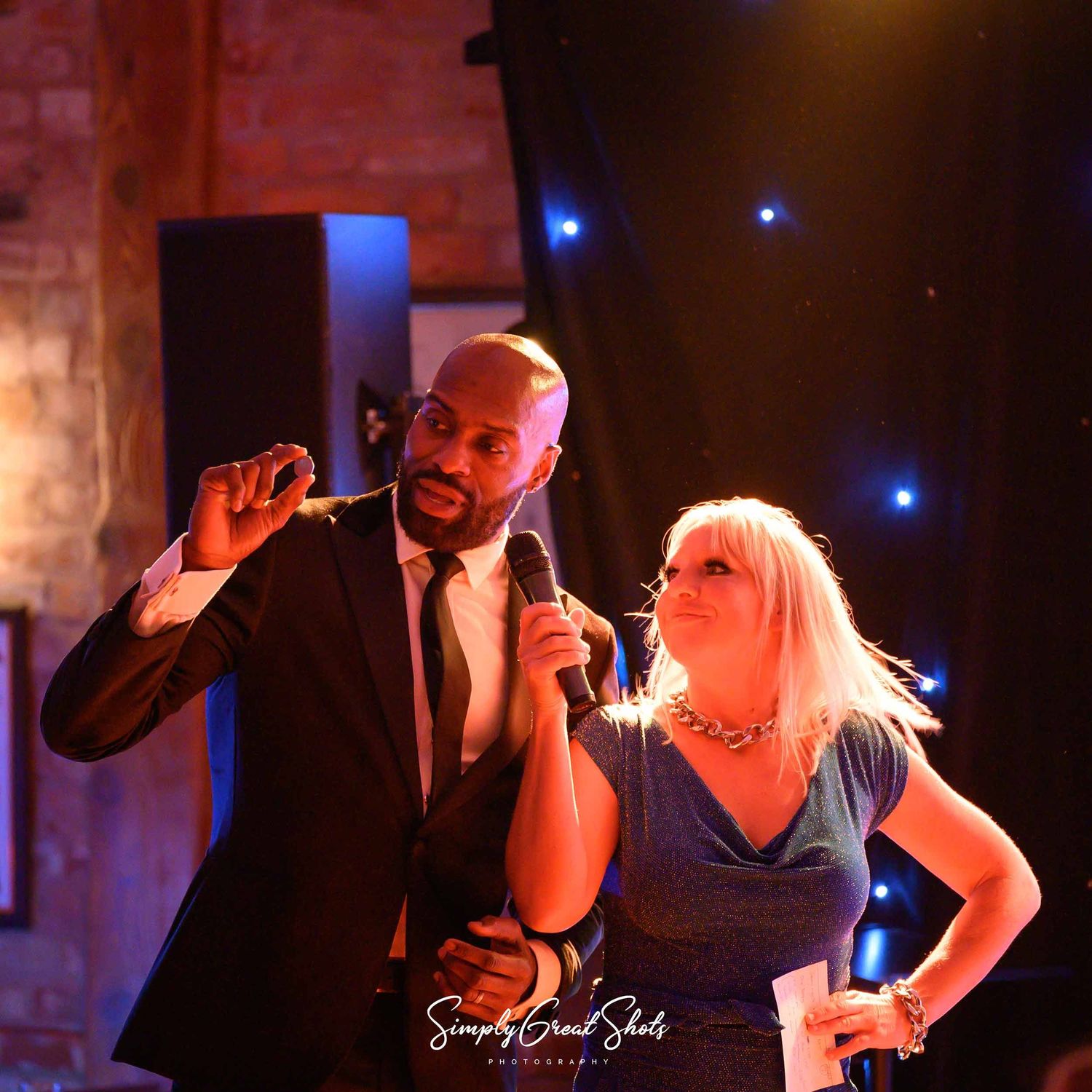 Two performers singing together with emotional expressions on a dimly lit stage with blue lighting and a brick wall backdrop.