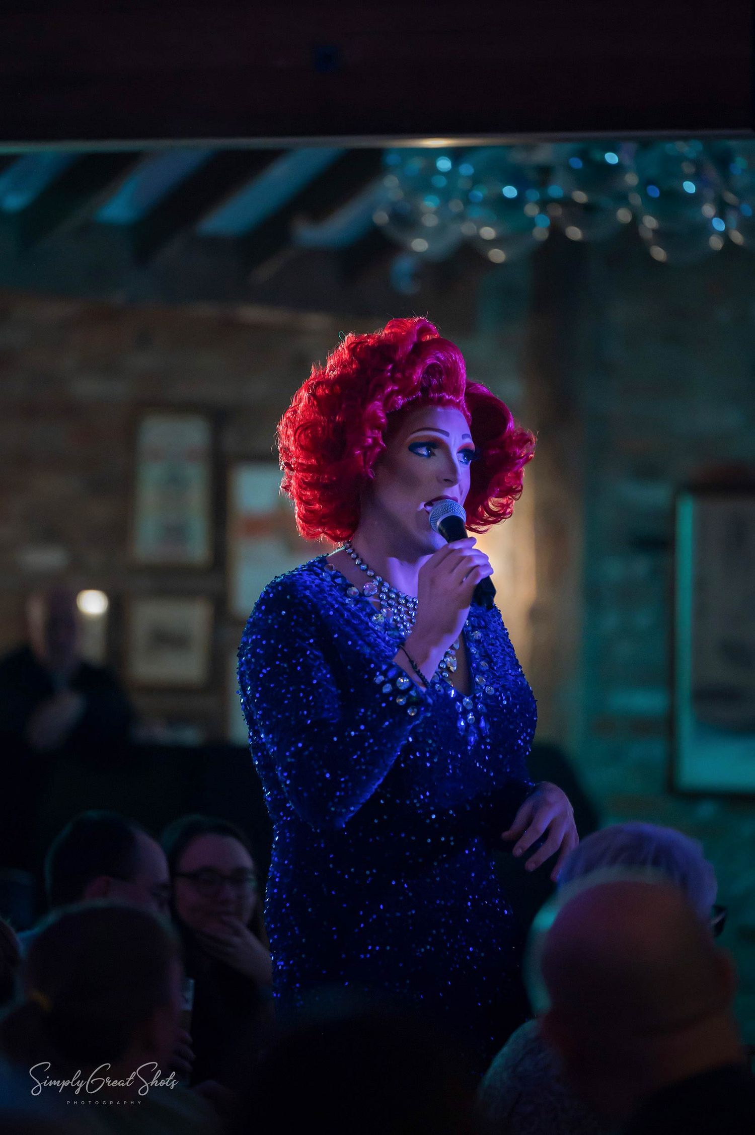 A performer in a sparkling blue sequined dress and bright red wig sings on stage in dramatic lighting.