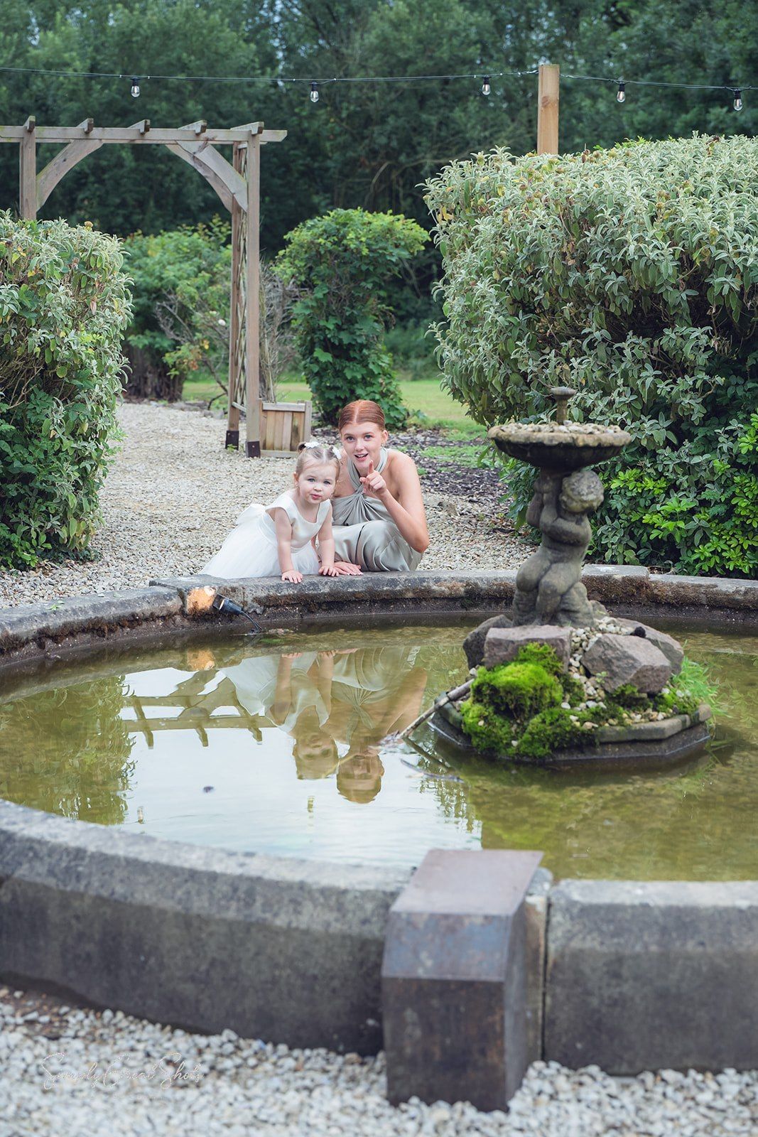 A bridesmaid and flower girl relax beside a peaceful garden pond with stone features and lush greenery in a tranquil outdoor setting.