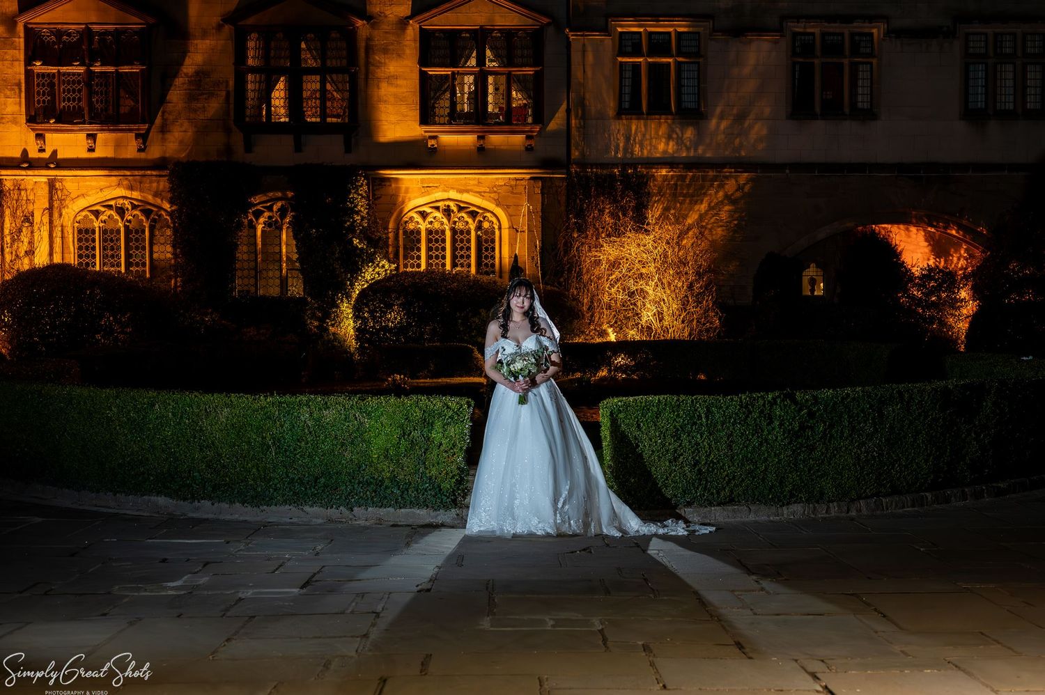 Bride Minky posing in her off-the-shoulder gown at night with the illuminated Coombe Abbey in the background.