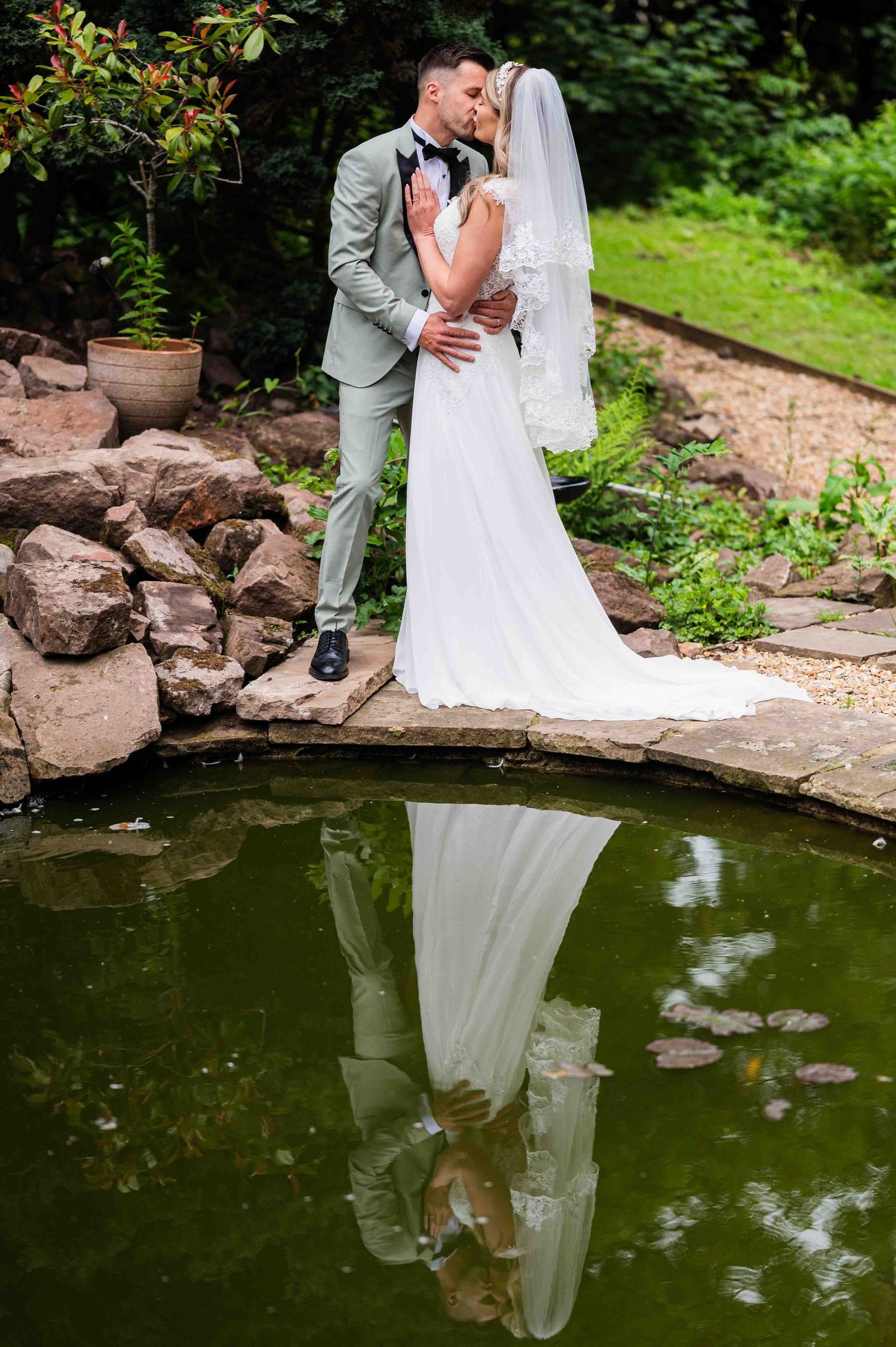 A bride and groom kiss in front of an ornamental pond, with their reflections visible in the water.