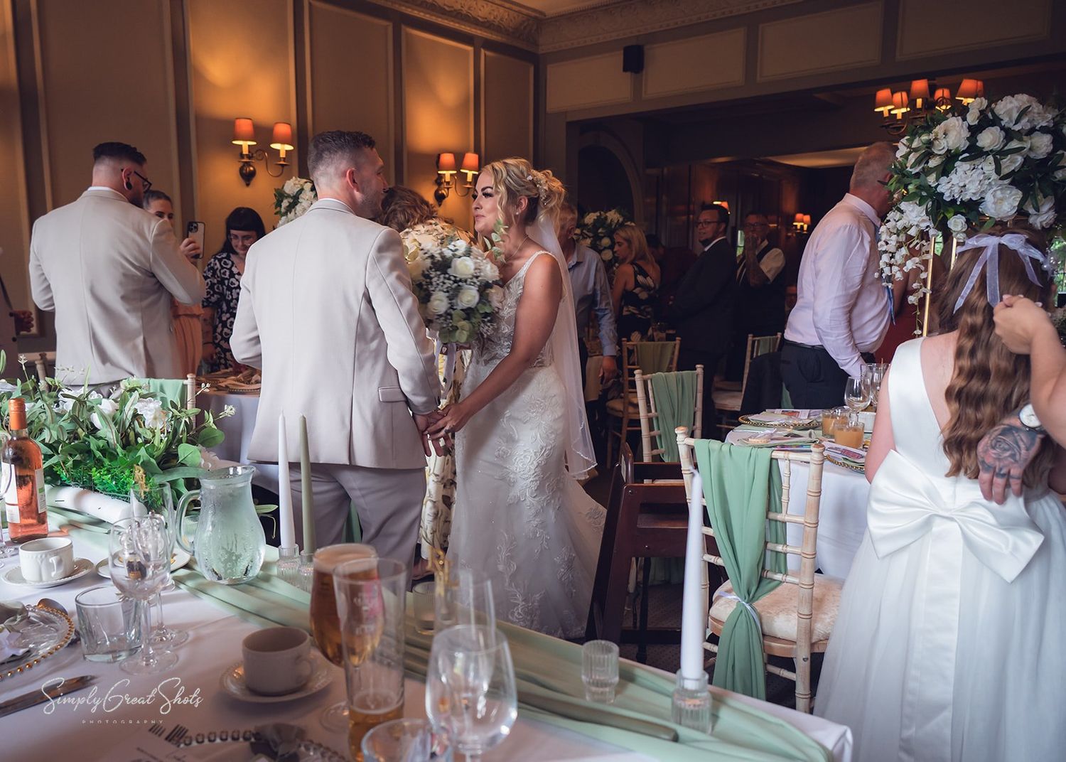 Wedding reception guests in light-coloured attire gather around an elegantly decorated dining area inside a banquet hall.