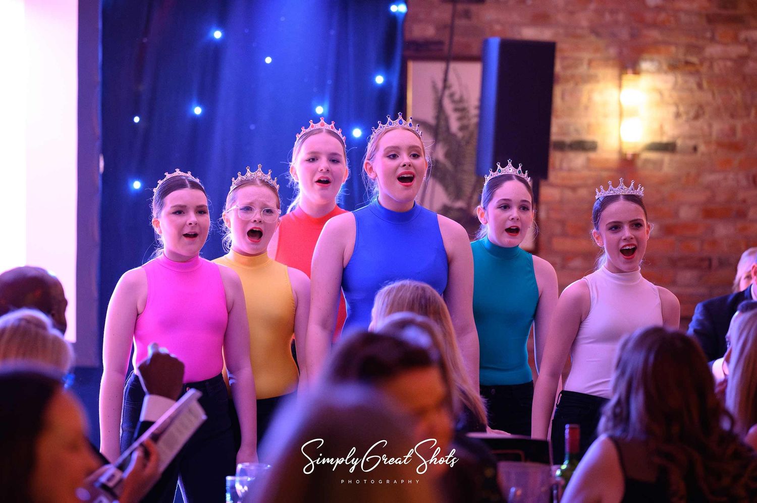 Performers wearing colorful leotards sing together on stage with blue lighting and brick wall backdrop.