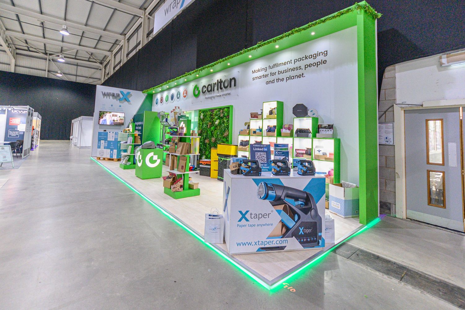 Modern trade show booth display with green walls showcasing electronics and gaming equipment in a spacious hall.