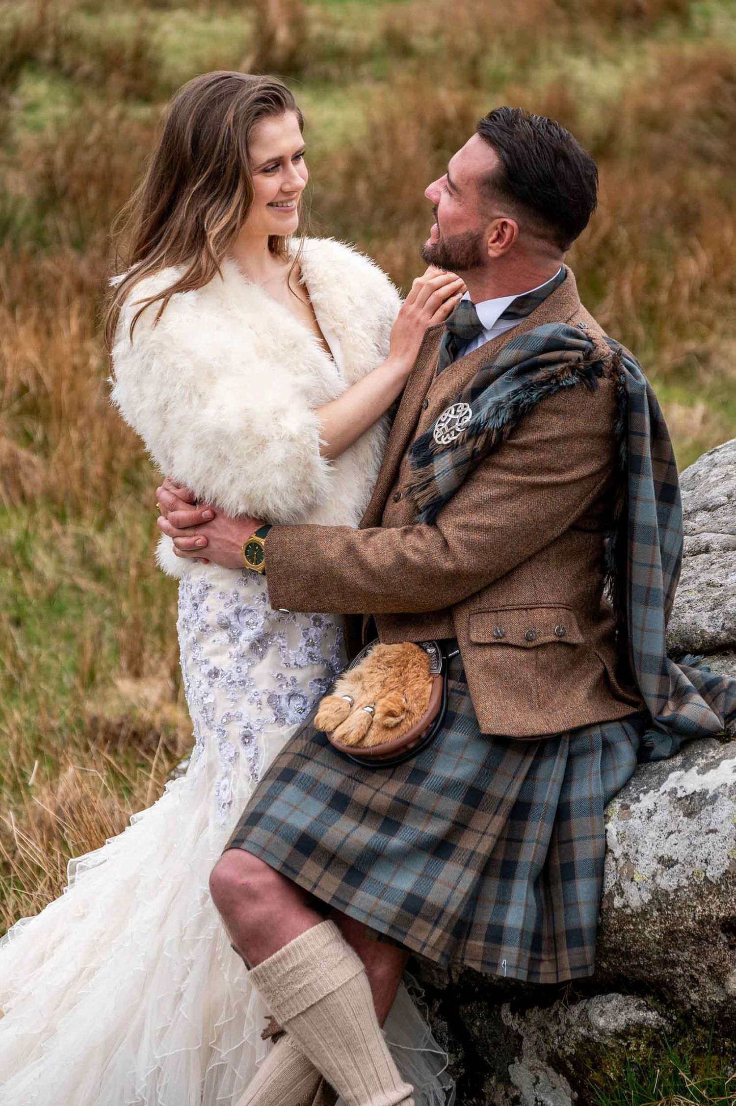 Scottish-style wedding photo with couple in traditional dress including tartan kilt and white fur shawl in rustic outdoor setting.